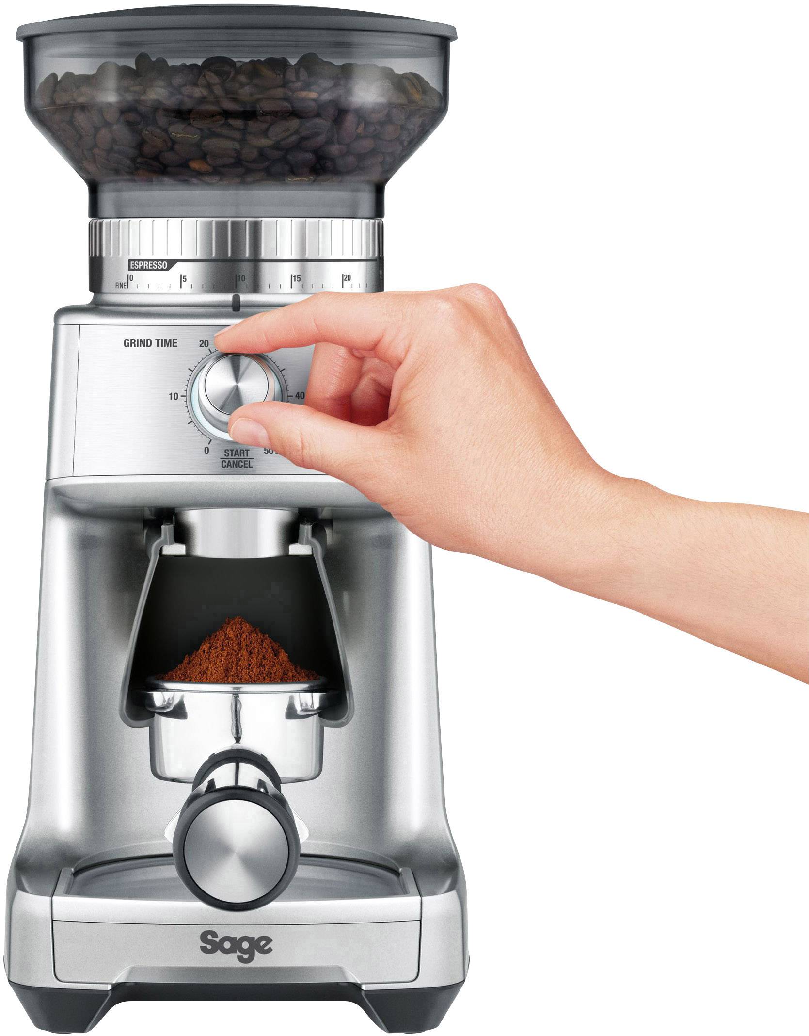 Une main tourne le réglage de la mouture sur un moulin à café argenté. Les grains de café sont dans le réservoir supérieur, et le café moulu se collecte en bas.