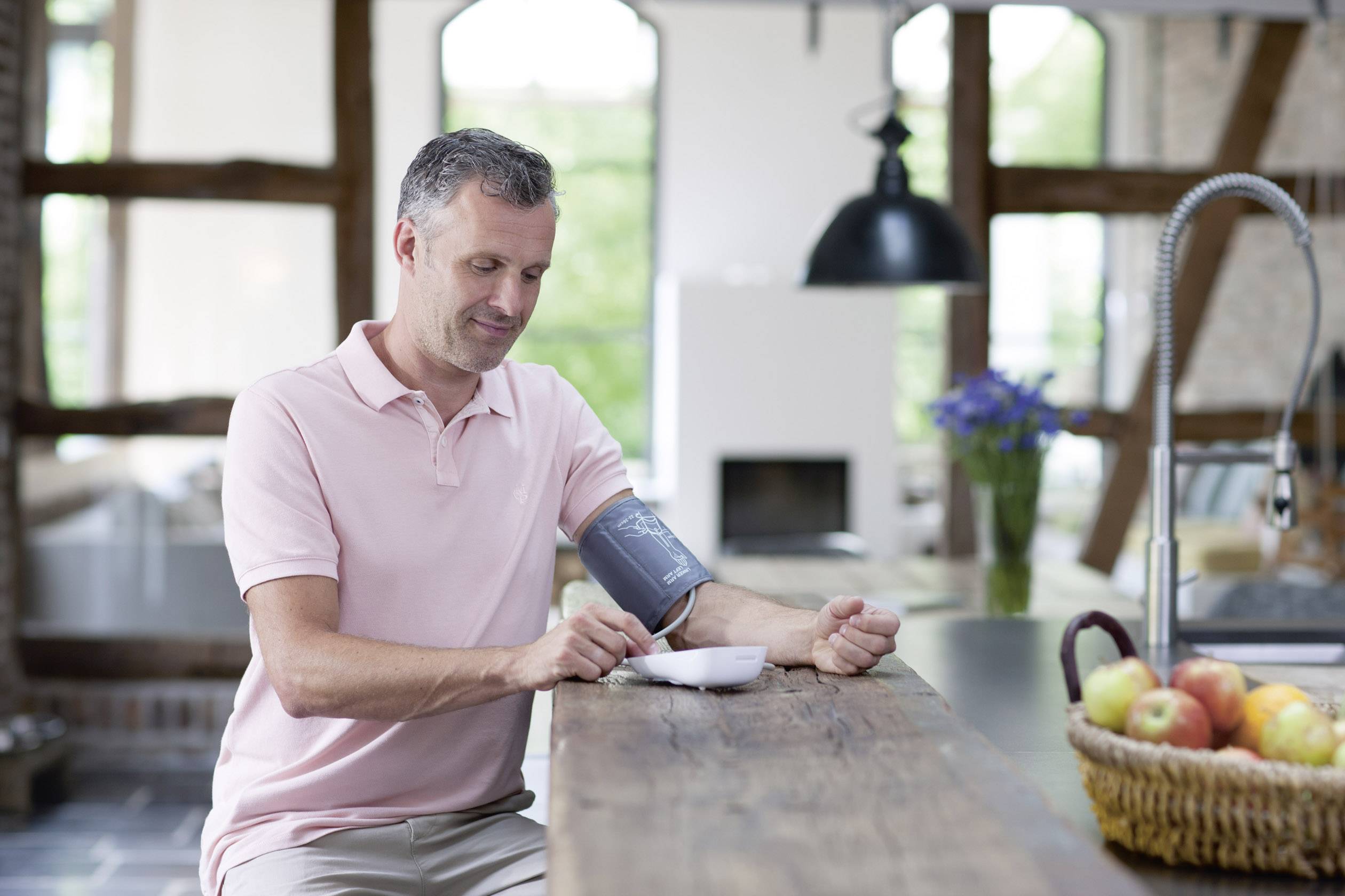 Un homme mesure sa tension artérielle dans une cuisine moderne. Un panier de fruits est posé sur la table.