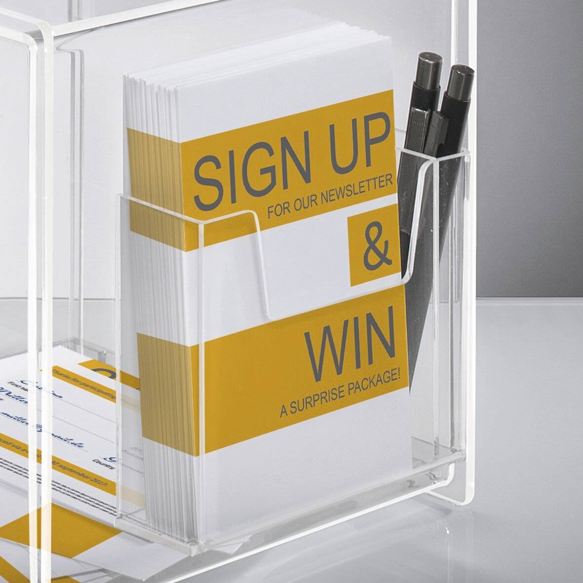 Pile de prospectus avec le texte « Inscrivez-vous à notre newsletter et gagnez un colis surprise ! » à côté de stylos, dans un support transparent sur un comptoir.