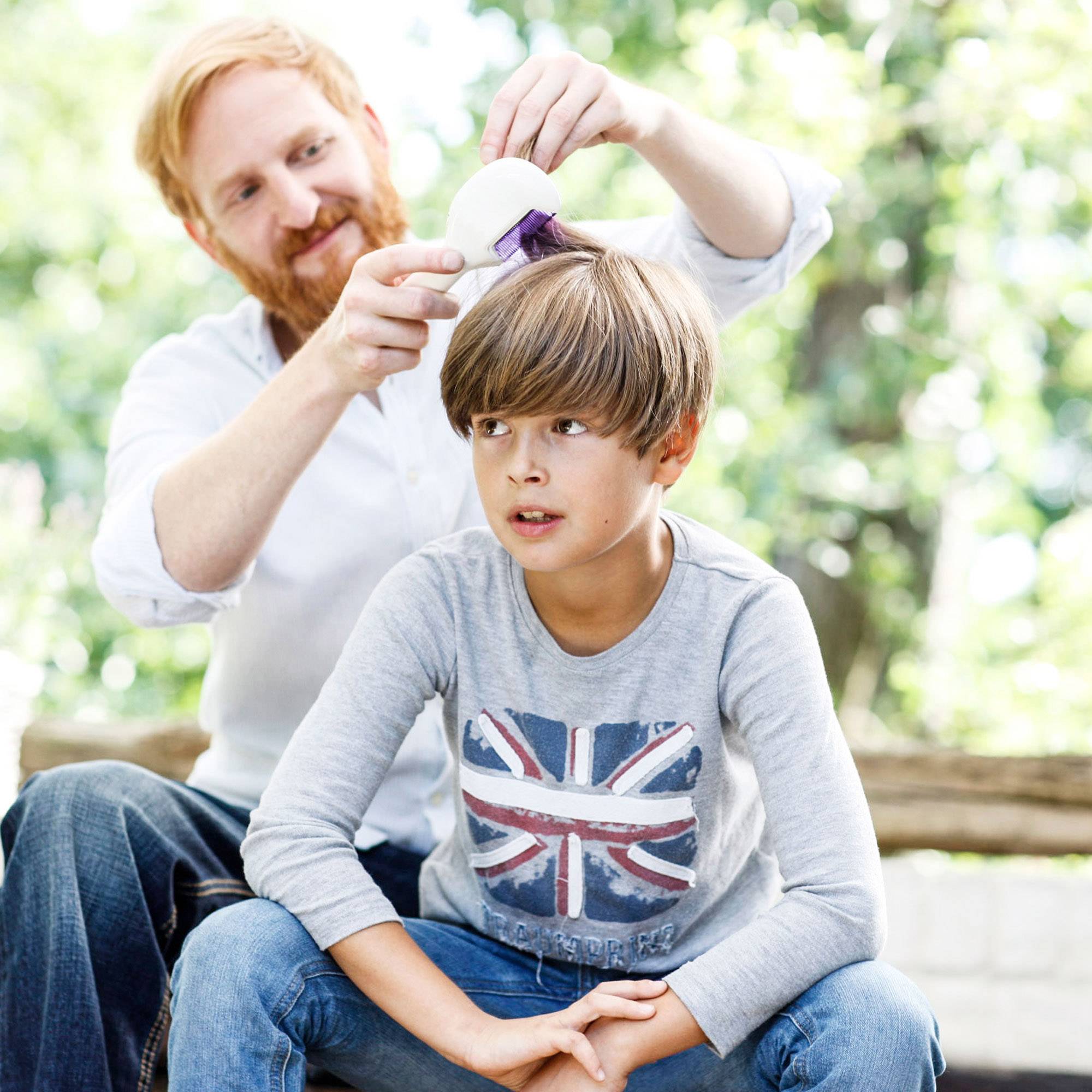 Un adulte peigne les cheveux d'un enfant avec un appareil électrique à l'extérieur. Tous deux sourient de manière détendue pendant le processus.