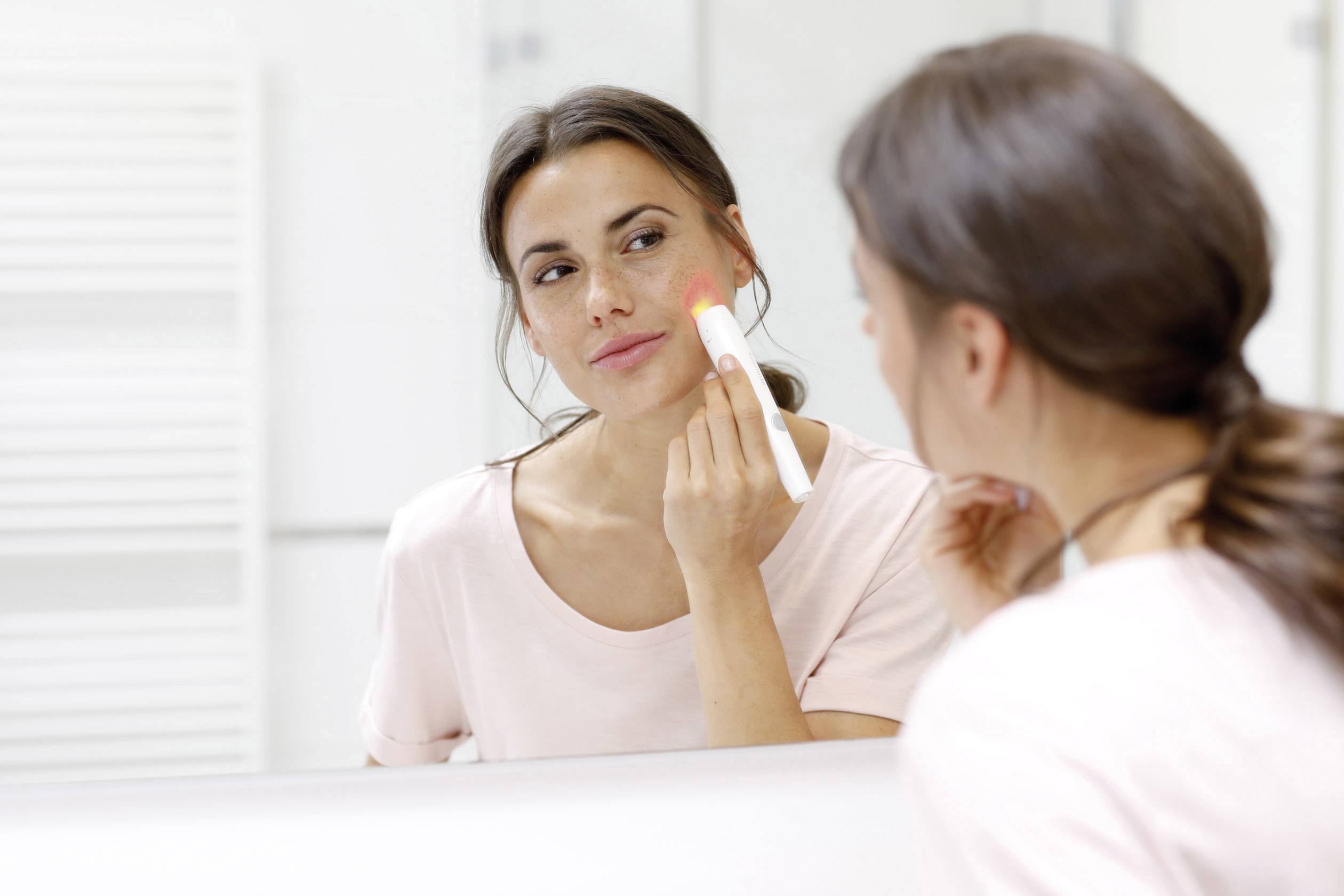 Une femme utilise un stick de maquillage devant un miroir. Elle a des cheveux foncés et porte un t-shirt clair. L'arrière-plan est flou.