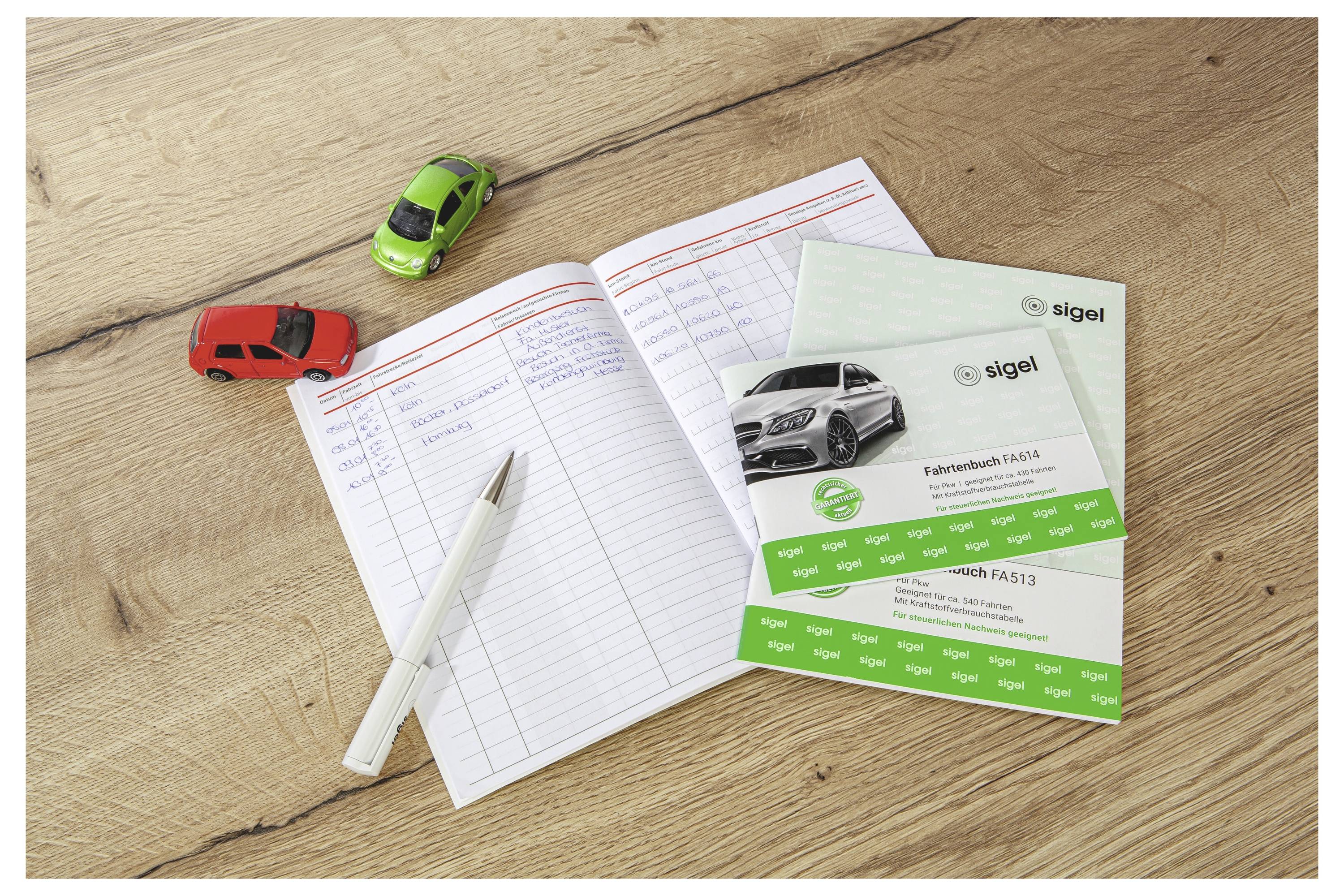 Carnet ouvert sur un bureau en bois à côté d'un stylo, deux voitures miniatures et des brochures automobiles Sigel, suggérant une planification ou une tenue de registres liés aux véhicules.