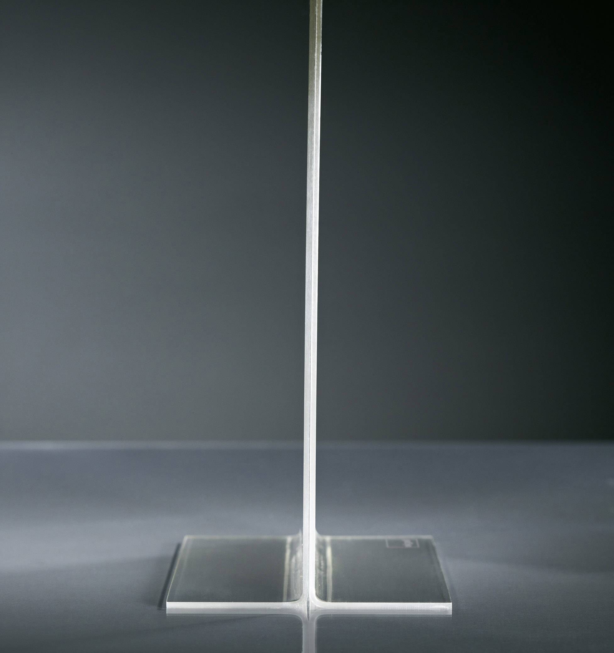 Objet acrylique transparent en forme de T, posé sur une surface réfléchissante devant un fond gris.