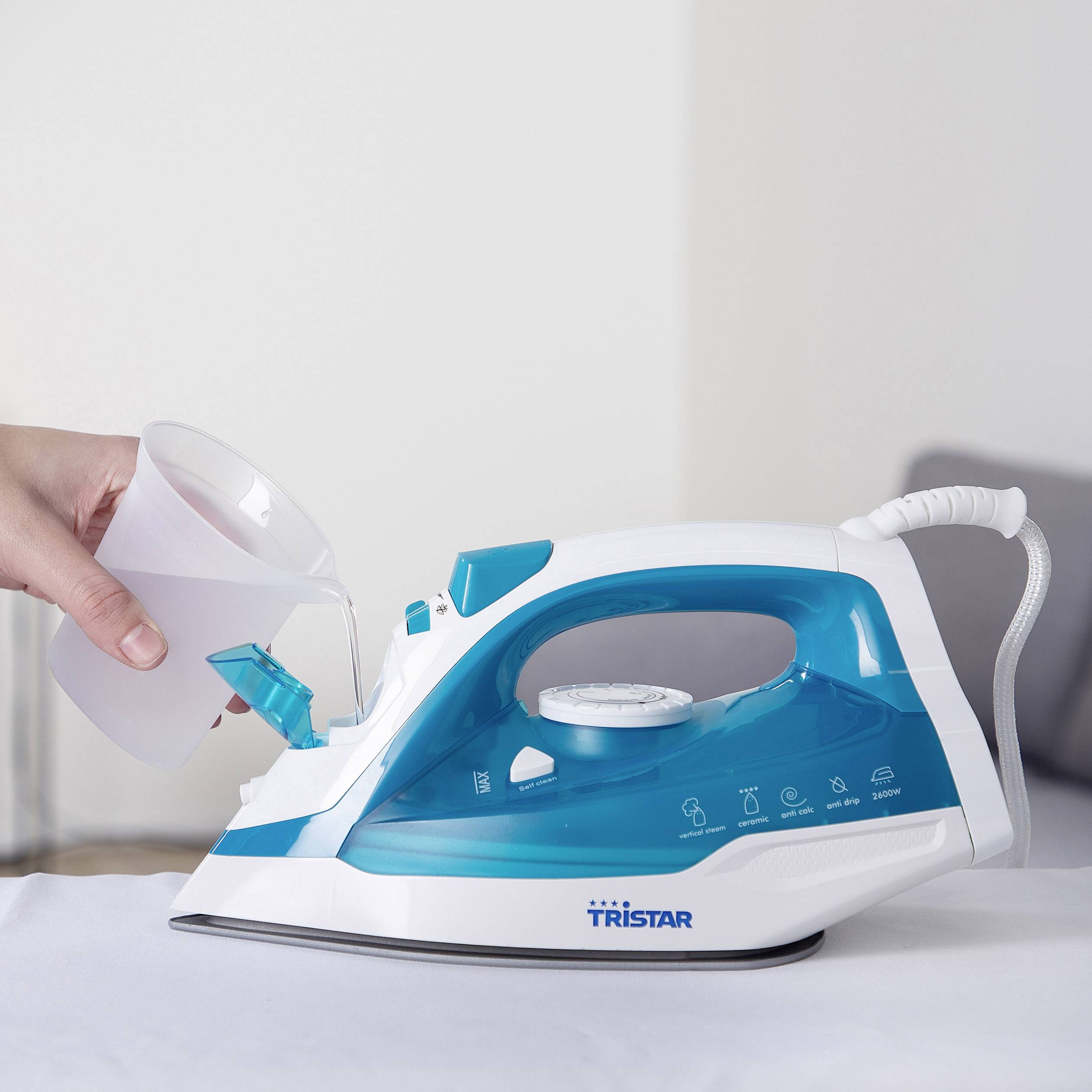 Une main remplit d'eau un fer à repasser de la marque Tristar à l'aide d'un gobelet, alors qu'il est posé sur une table.