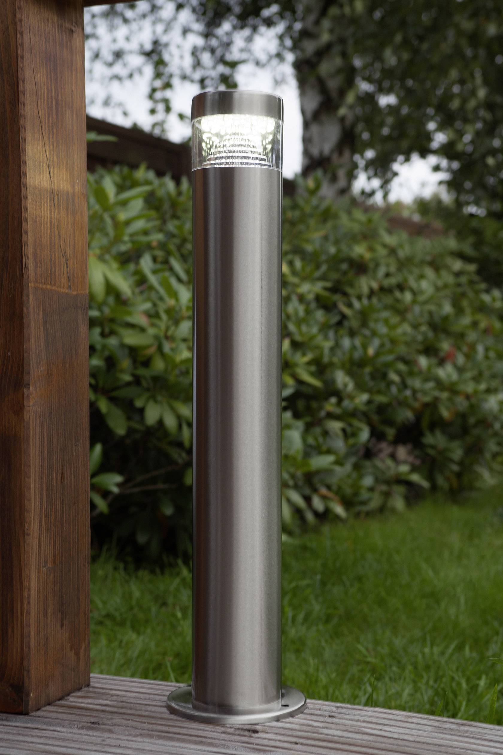 Lampe solaire en acier inoxydable posée sur une terrasse en bois, entourée de plantes.