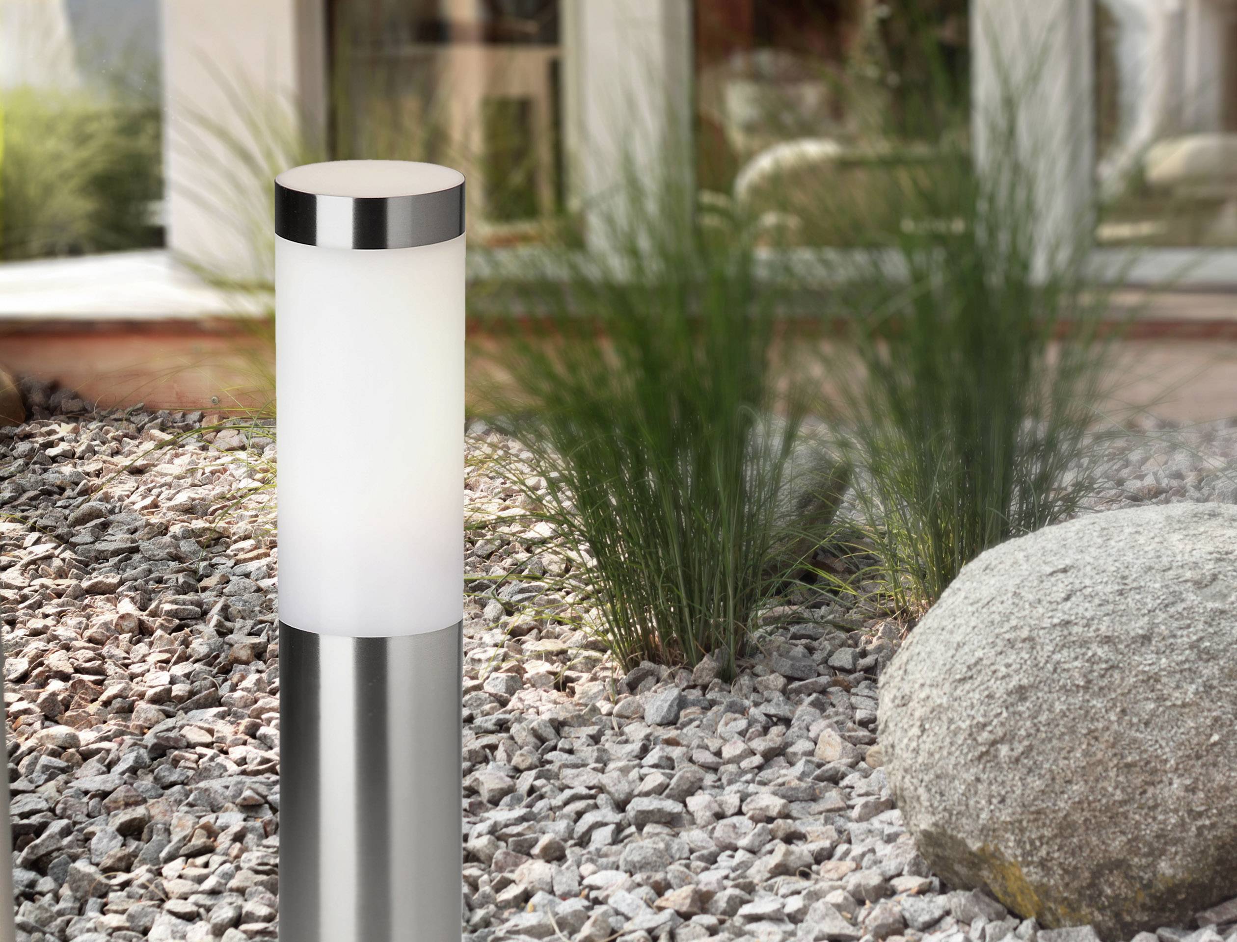 Un lampadaire de jardin moderne en acier inoxydable se dresse sur un sol de gravier, entouré d'herbes vertes et d'une grande pierre. Un bâtiment est visible en arrière-plan.