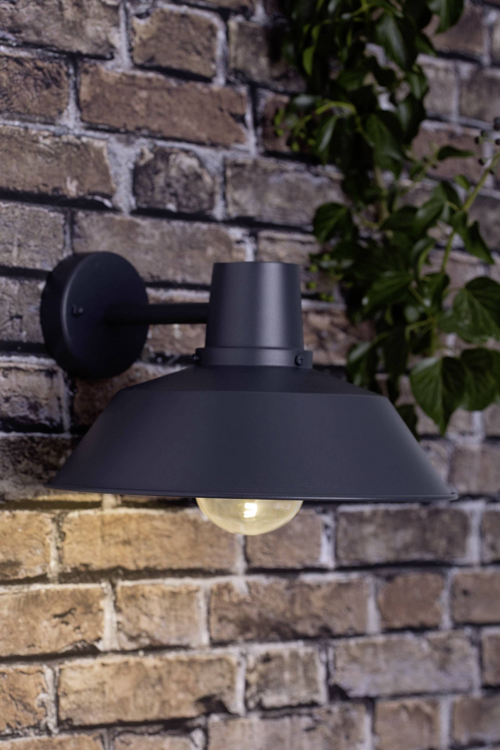 Lampe extérieure montée sur un mur de briques, avec un abat-jour noir, illuminant légèrement les alentours. Une plante verte à proximité.