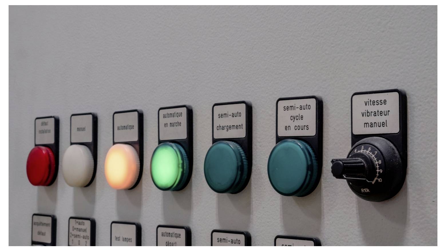 Rangée de boutons de commande industriels et de sélecteurs sur un panneau mural, avec un bouton rouge, un blanc, un ambre illuminé, un vert allumé, et des étiquettes en français telles que 'semi-auto chargement' et 'vitesse vibrateur manuel'.