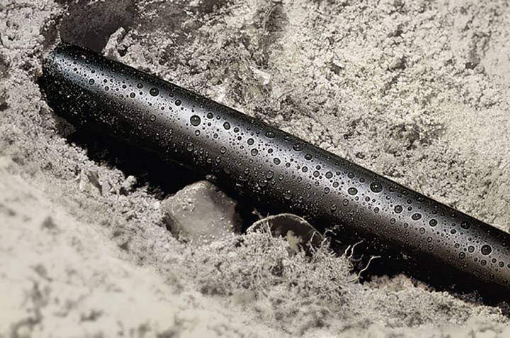 Un tuyau noir est à moitié enterré dans le sol sablonneux et recouvert de petites gouttelettes d'eau.
