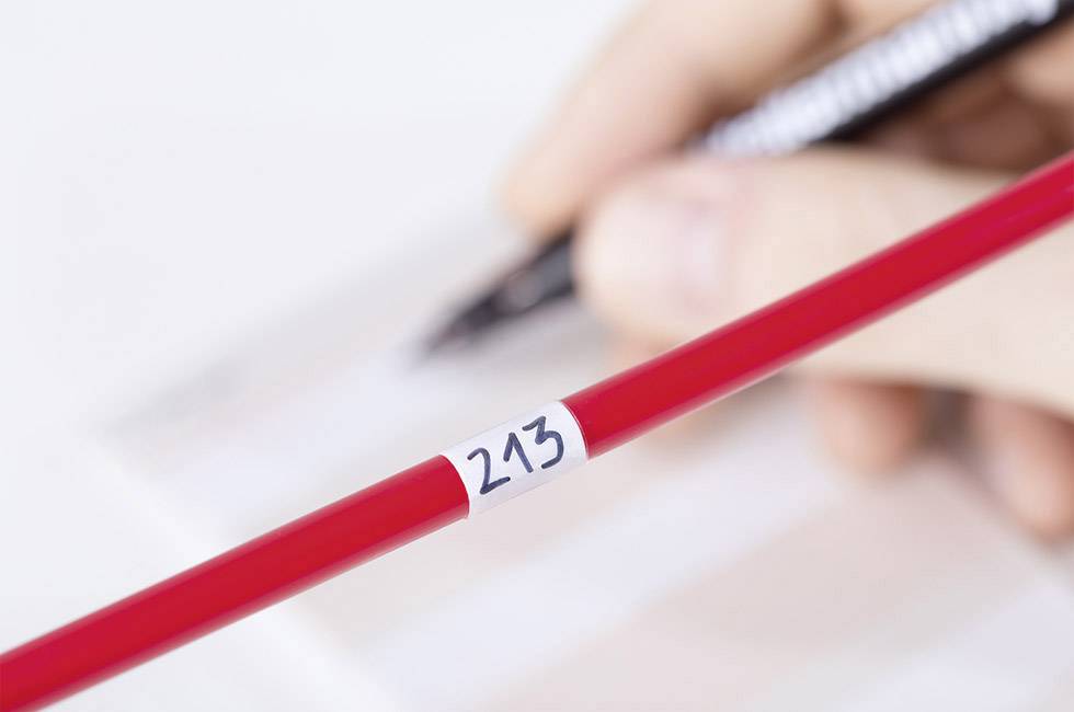 Une main écrit avec un stylo noir sur du papier. Au premier plan, un crayon rouge portant le numéro '243' est posé.