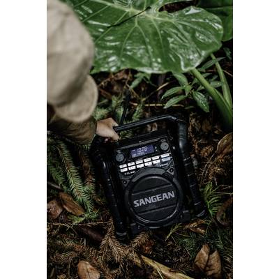 Une personne tient un haut-parleur radio portable robuste dans la jungle. La radio affiche '10:08'. Le joueur est positionné sur le sol forestier.