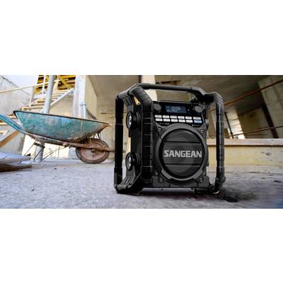 Radio portable de chantier posé sur un sol de construction, à côté d'une brouette. Robuste et fonctionnel pour écouter de la musique ou des informations.
