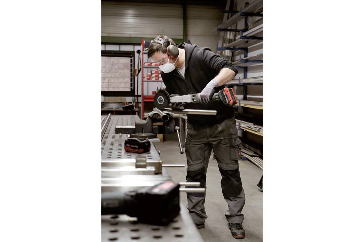 Un ouvrier en tenue de protection meule du métal dans un atelier. Il utilise une meuleuse électrique sur une table de travail.