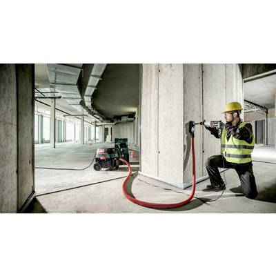 Un ouvrier en équipement de protection utilise un outil électrique pour percer un trou dans un mur en béton. Une machine d'aspiration des poussières est branchée.