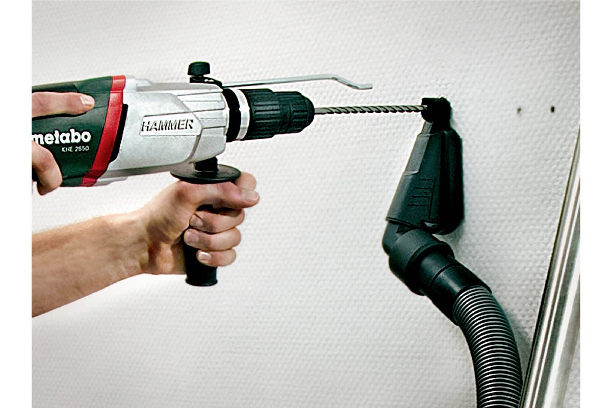 Une main tient une perceuse qui fore un trou dans un mur blanc. Un embout d'aspirateur récupère directement la poussière de forage au niveau du trou.