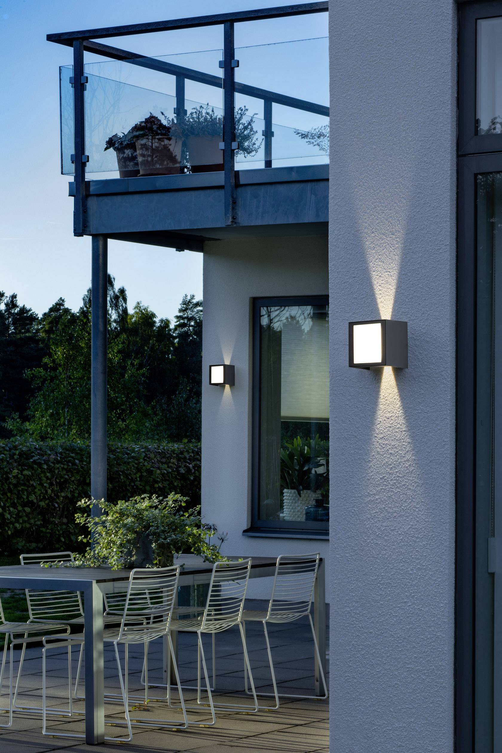 Une terrasse moderne au crépuscule avec une balustrade en verre, une table et des chaises éclairées ; des appliques murales illuminent le mur de la maison.