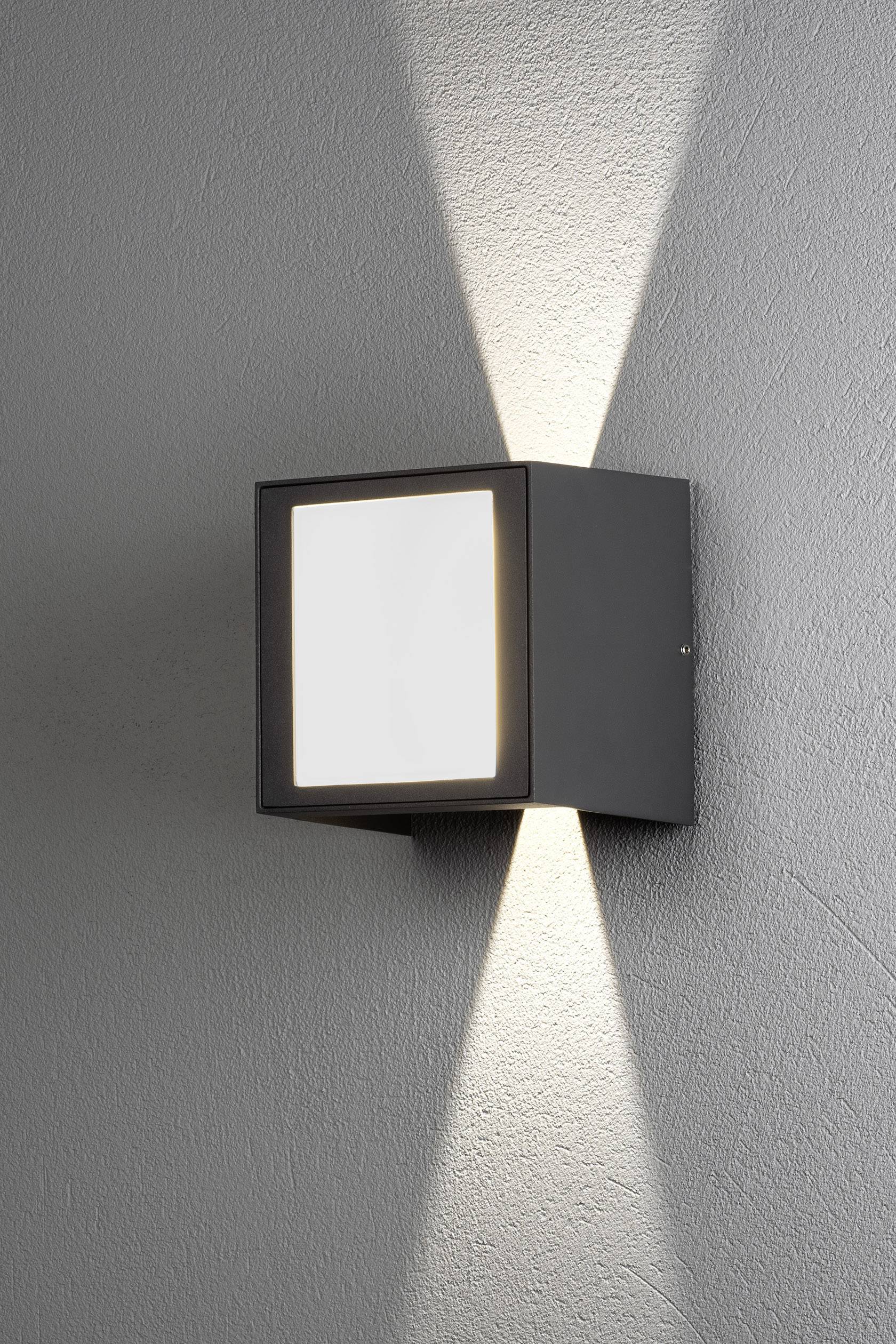 Applique murale au design carré projetant des rayons lumineux vers le haut et le bas sur un mur gris. Aspect moderne et minimaliste.