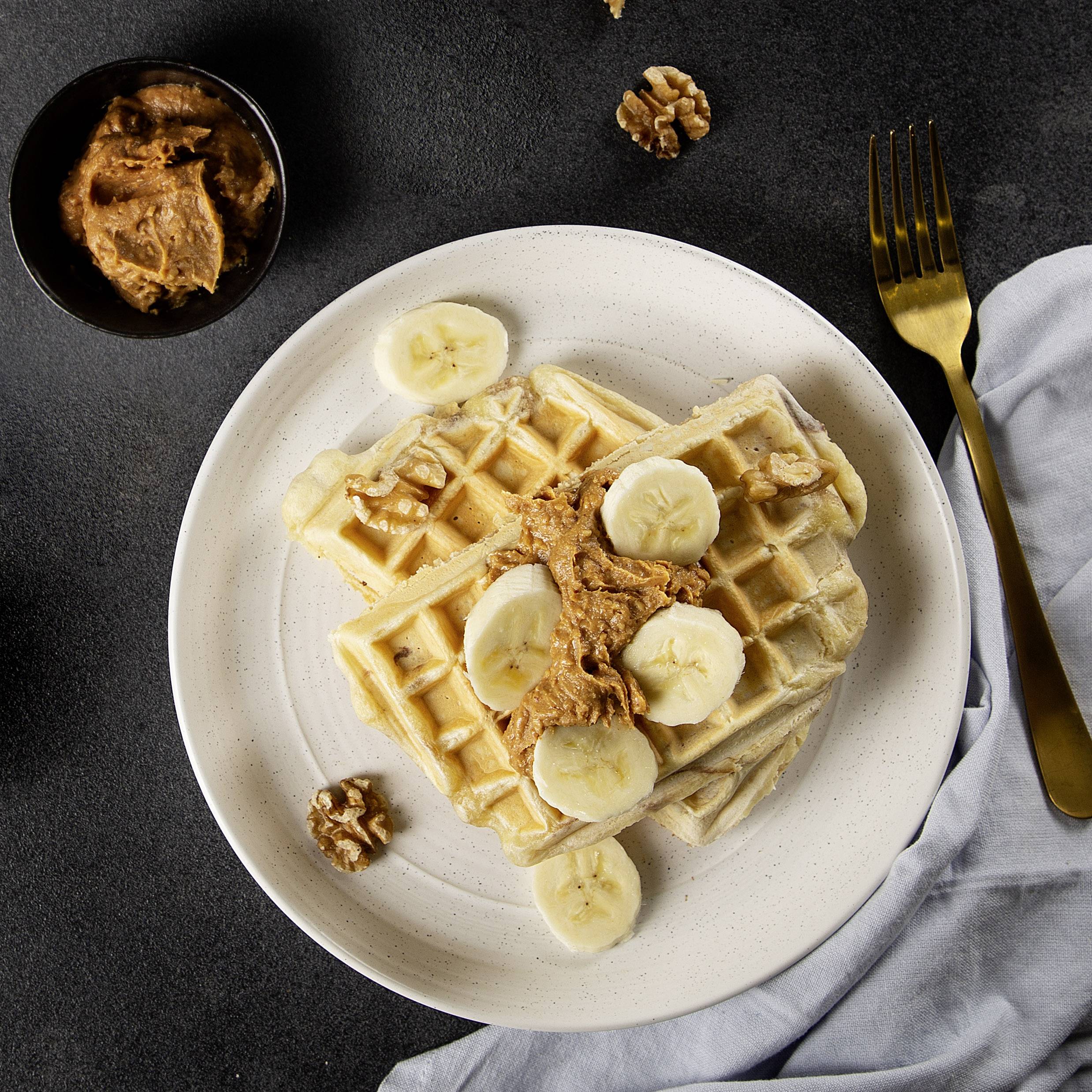 Une assiette avec deux gaufres, garnies de beurre de cacahuète, de tranches de banane et de noix. À gauche, un bol de beurre de cacahuète.