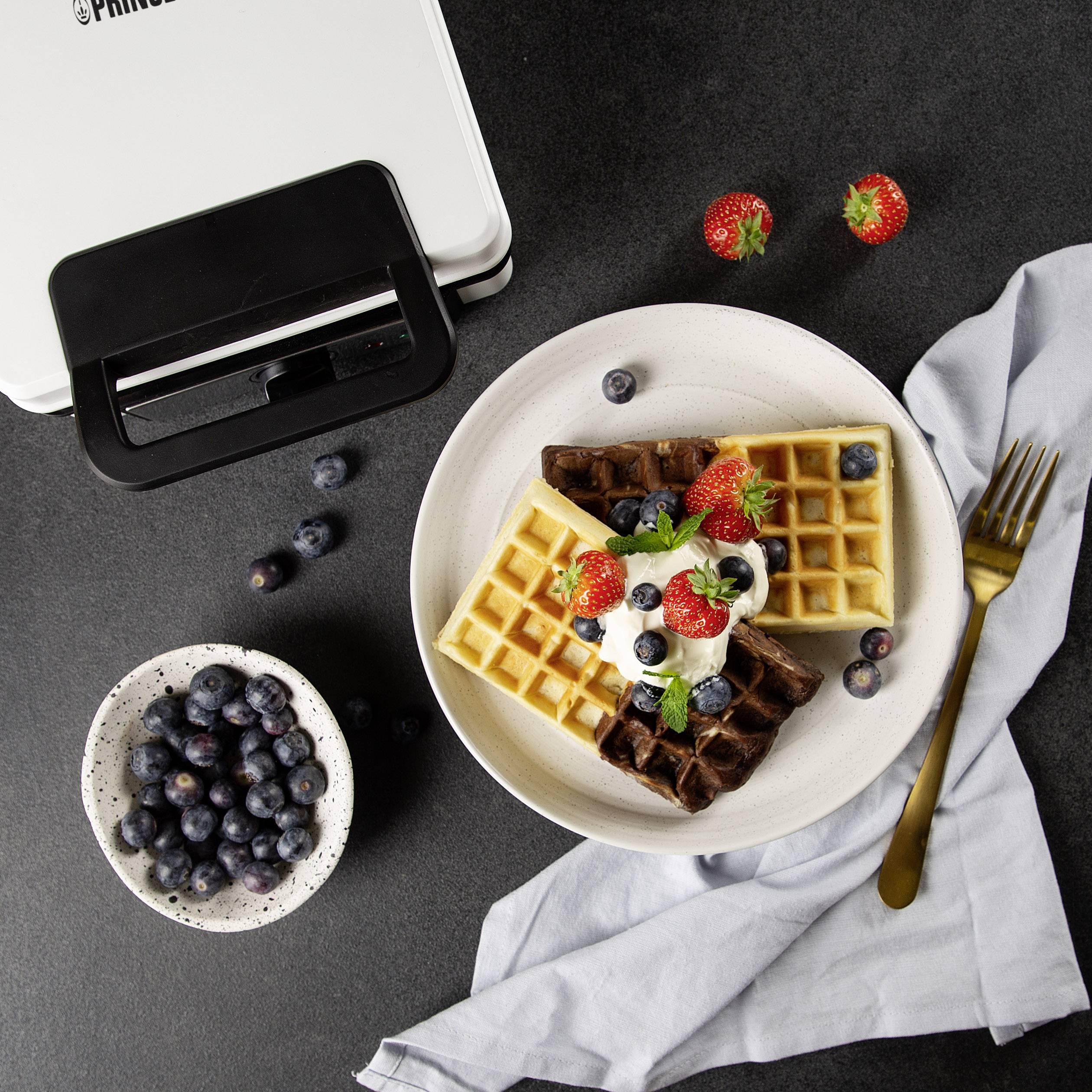 Une assiette de gaufres garnies de fraises, de myrtilles et de crème. À côté, des myrtilles et des fraises fraîches ainsi qu'un gaufrier sont disposés.