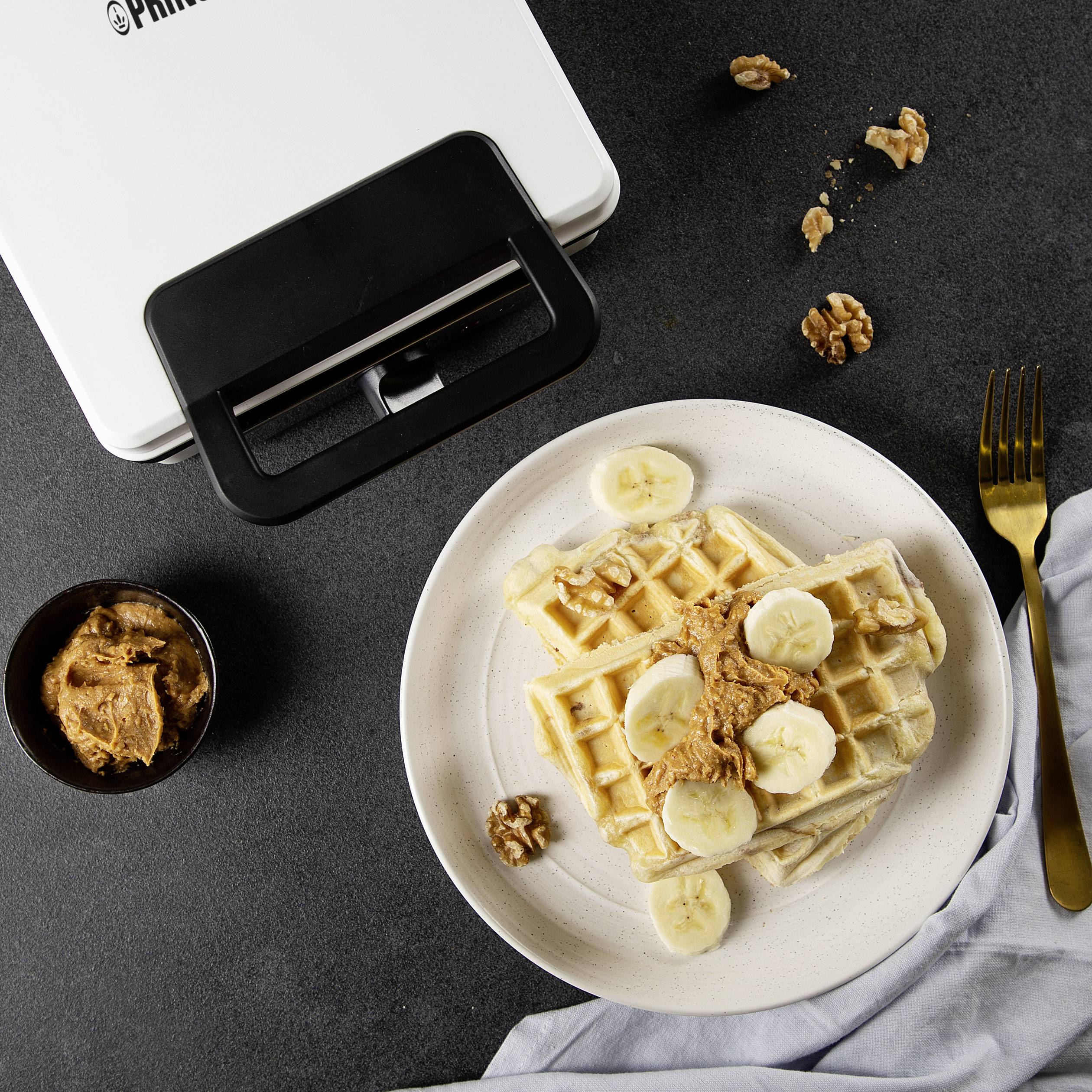 Une assiette de gaufres garnies de tranches de banane et de beurre de cacahuète. À côté, une fourchette et un gaufrier en arrière-plan.