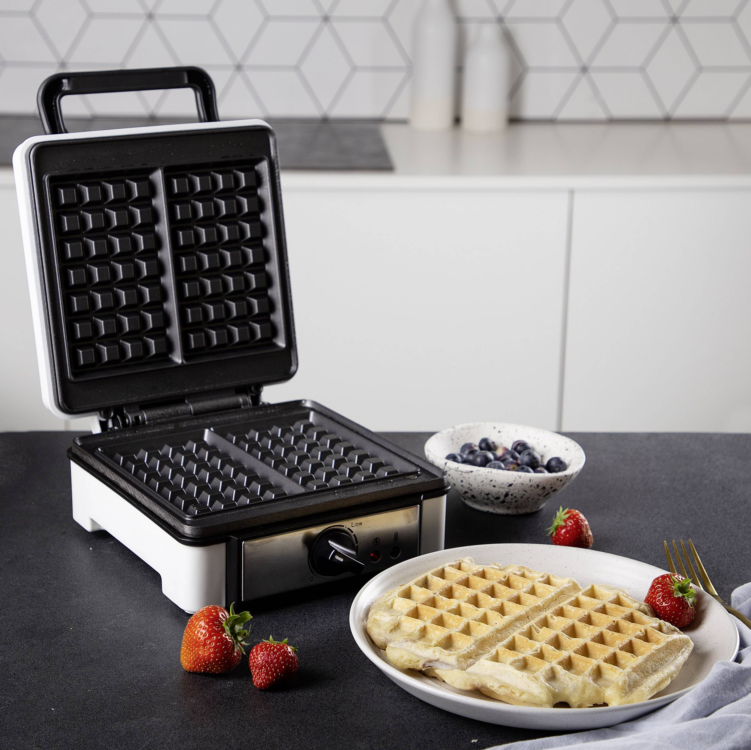 Un gaufrier ouvert sur un comptoir de cuisine avec deux gaufres prêtes dans une assiette. À côté, des bols de fruits rouges sont disposés.