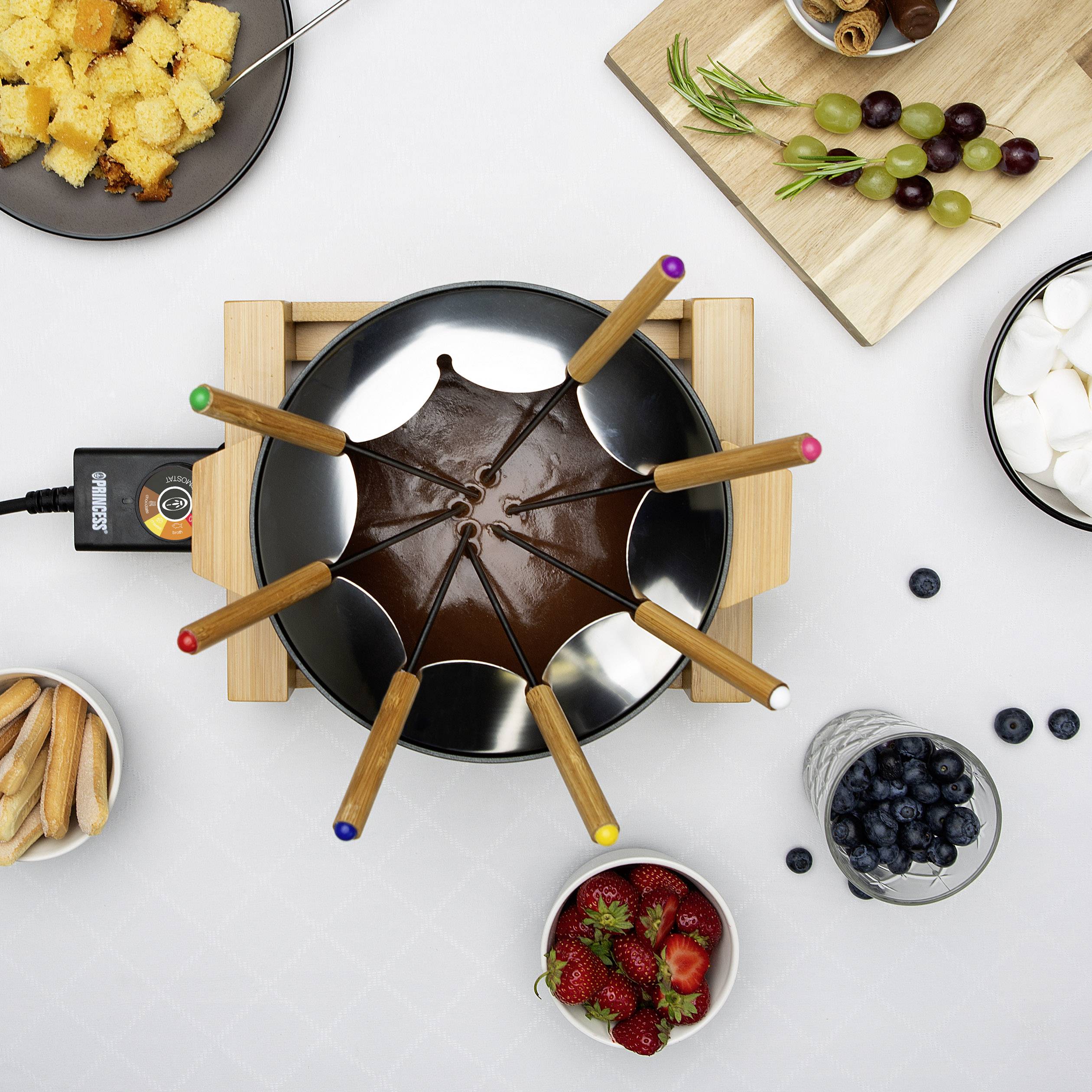 Une fondue au chocolat est entourée de fruits, de biscuits, de pain et de guimauves sur une table, prête à être trempée.