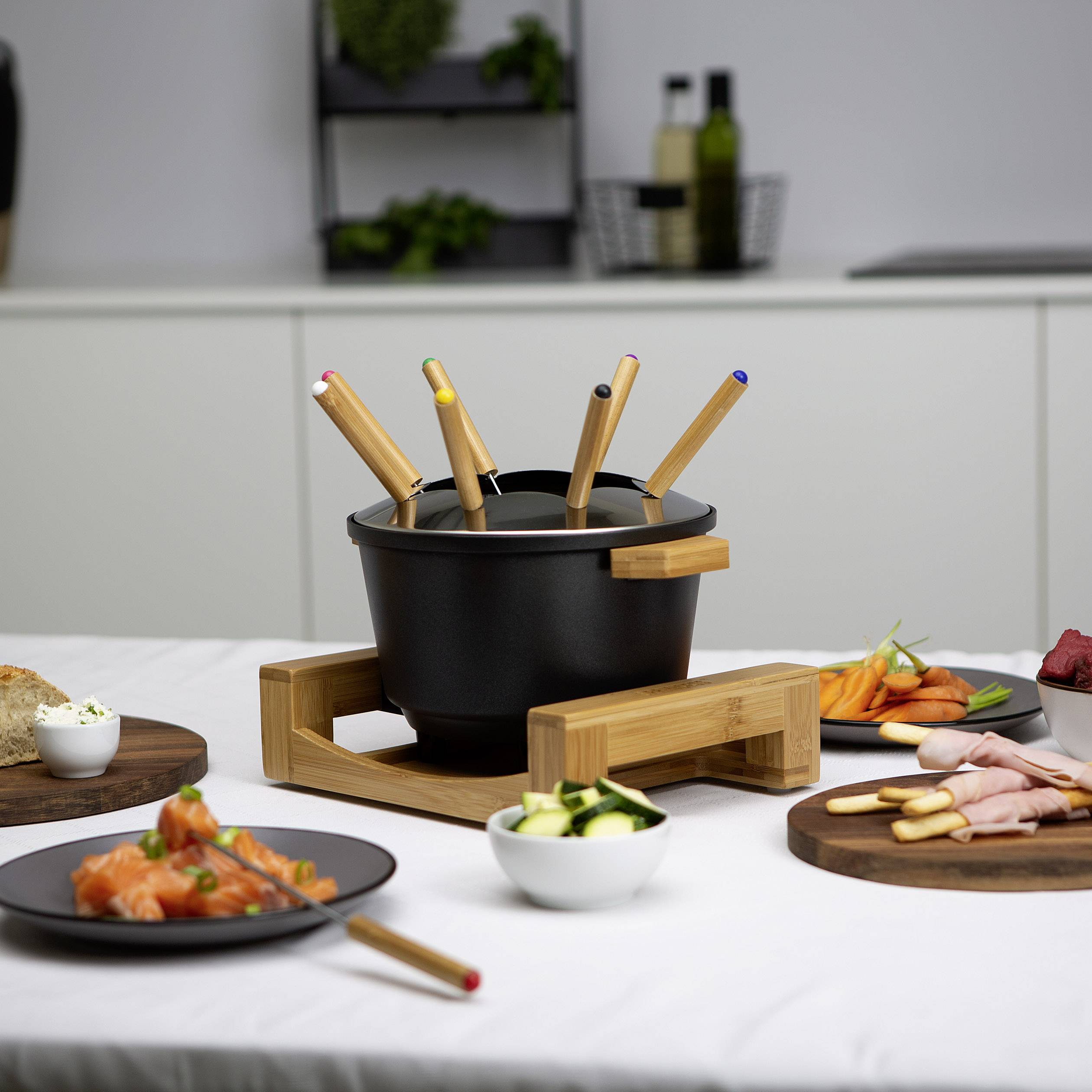 Pot de fondue au fromage, entouré d'assiettes de saumon, de légumes et de pain sur une table dans une cuisine moderne.