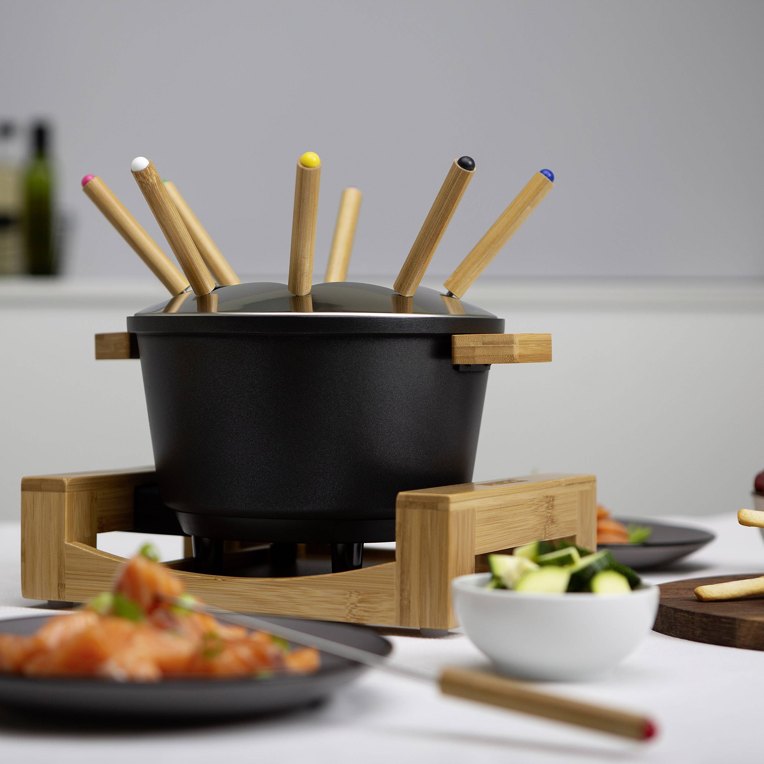 Marmite à fondue noire avec des fourchettes colorées, entourée de plats de légumes et de saumon sur une table dressée.