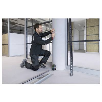 Une personne s'agenouille tout en mesurant une colonne cylindrique dans un espace intérieur moderne et inachevé, avec des outils de construction à proximité.