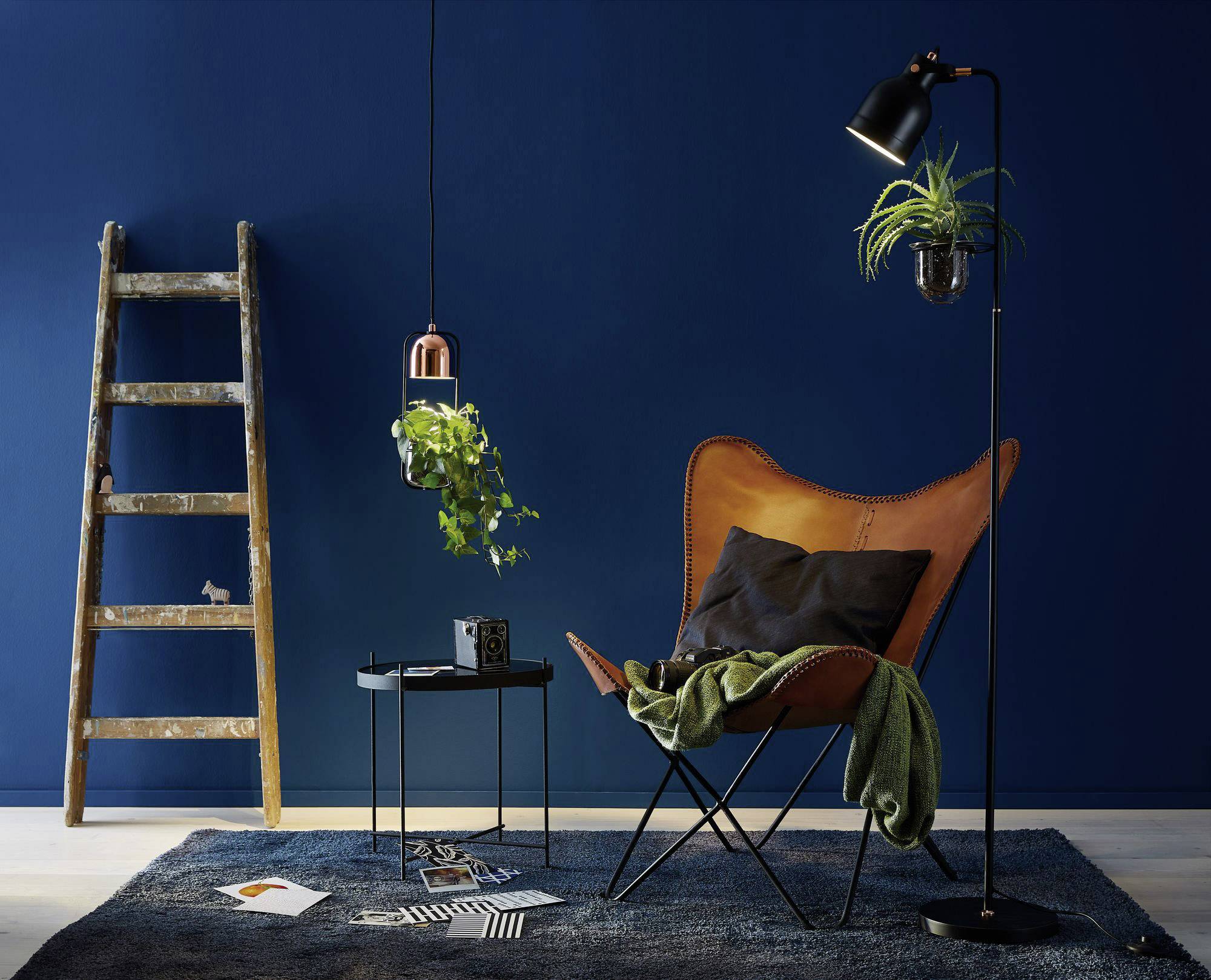 Un espace chaleureux avec un fauteuil en cuir marron, une couverture verte, un lampadaire avec une plante suspendue, une échelle en bois et un mur peint en bleu marine.
