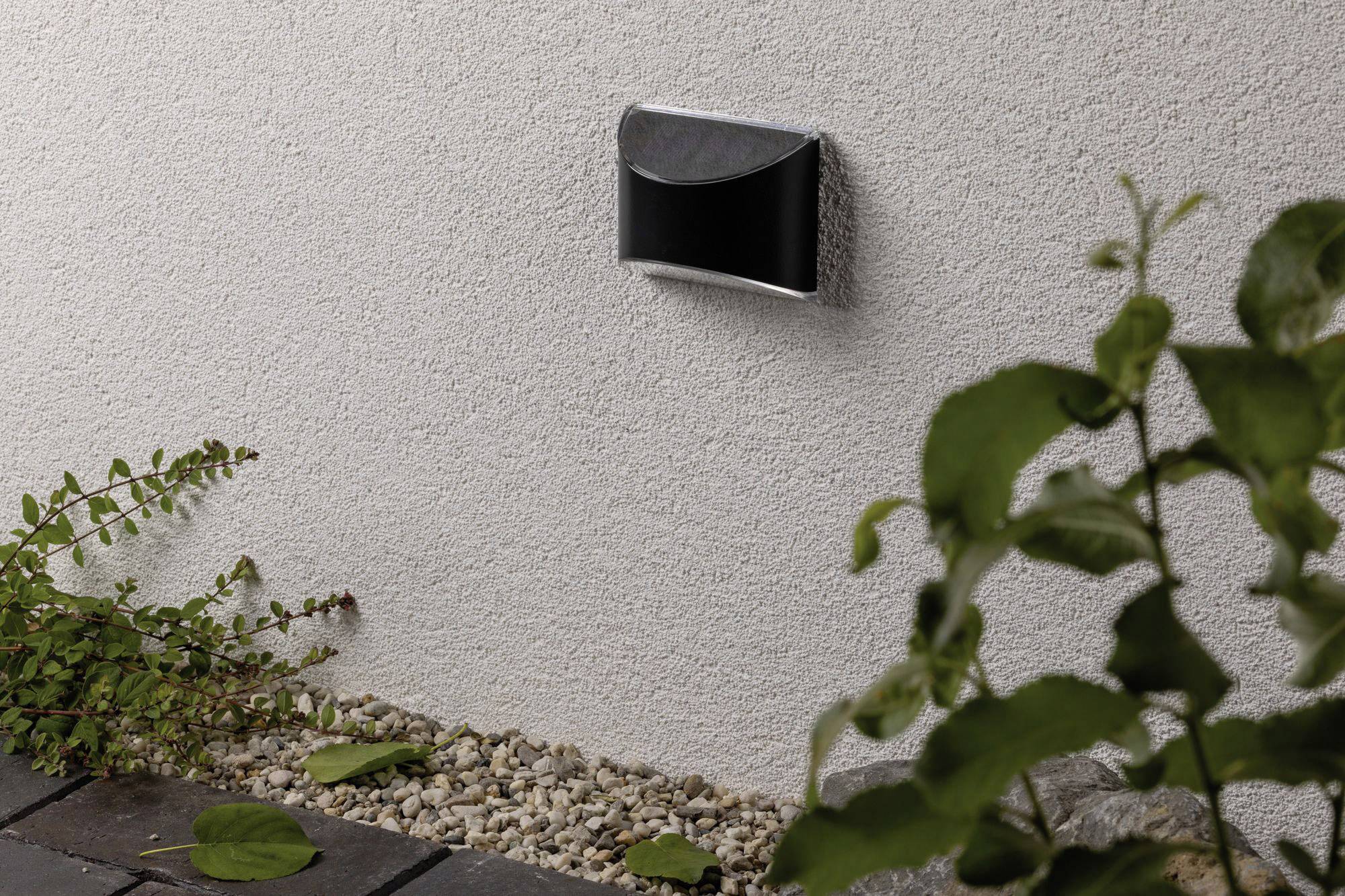 Un petit luminaire mural extérieur sur un mur blanc, entouré de plantes et de galets. Le luminaire est noir et de forme semi-circulaire.