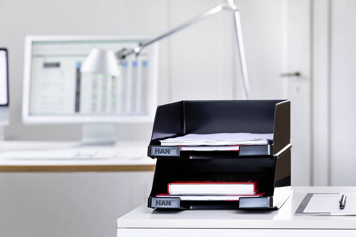 Bureau avec des plateaux empilables, remplis de documents, au premier plan ; à l'arrière-plan, un ordinateur sur un bureau.