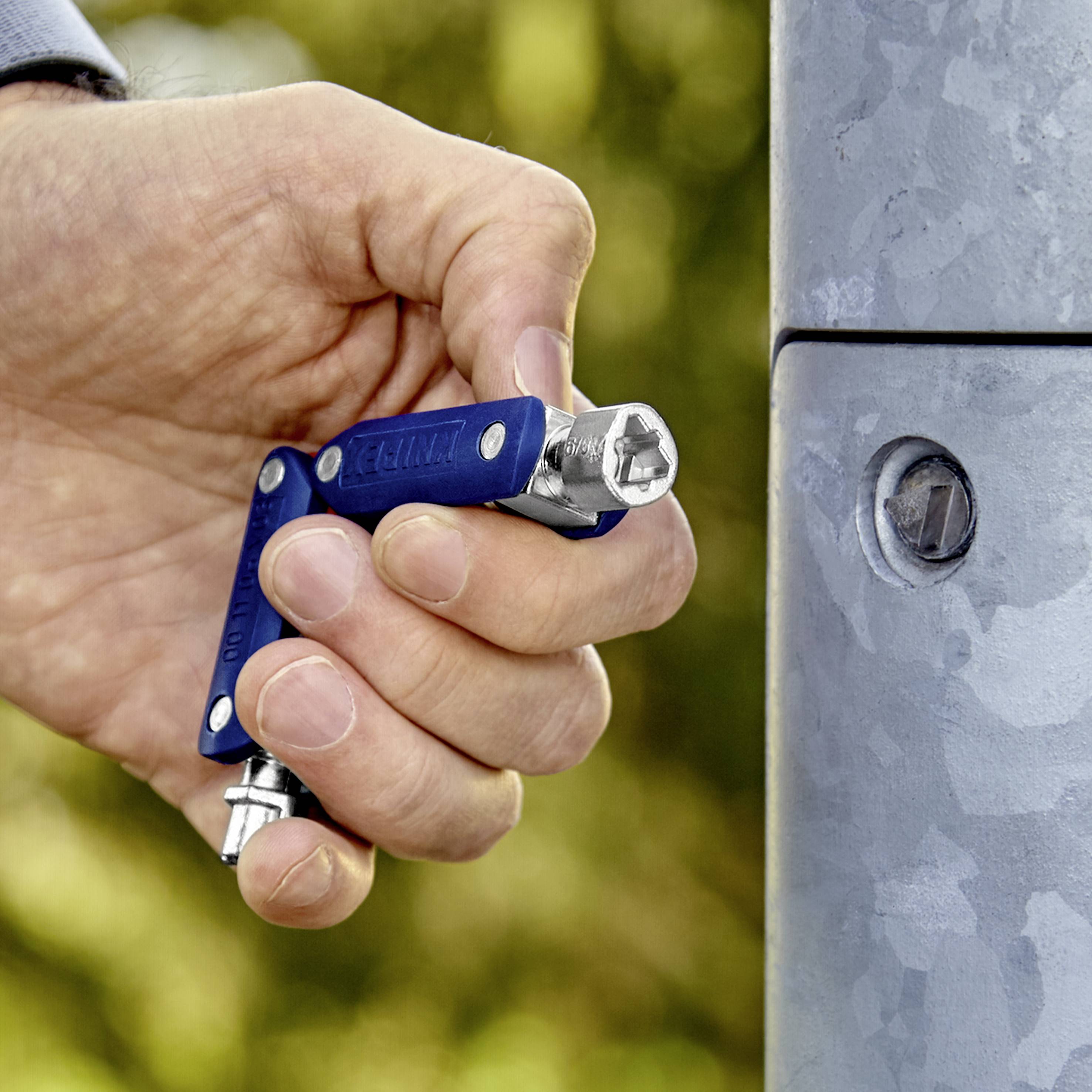 Une main tient un multi-outil bleu et serre une vis sur un poteau métallique. L'arrière-plan est flou et montre de la végétation verte.