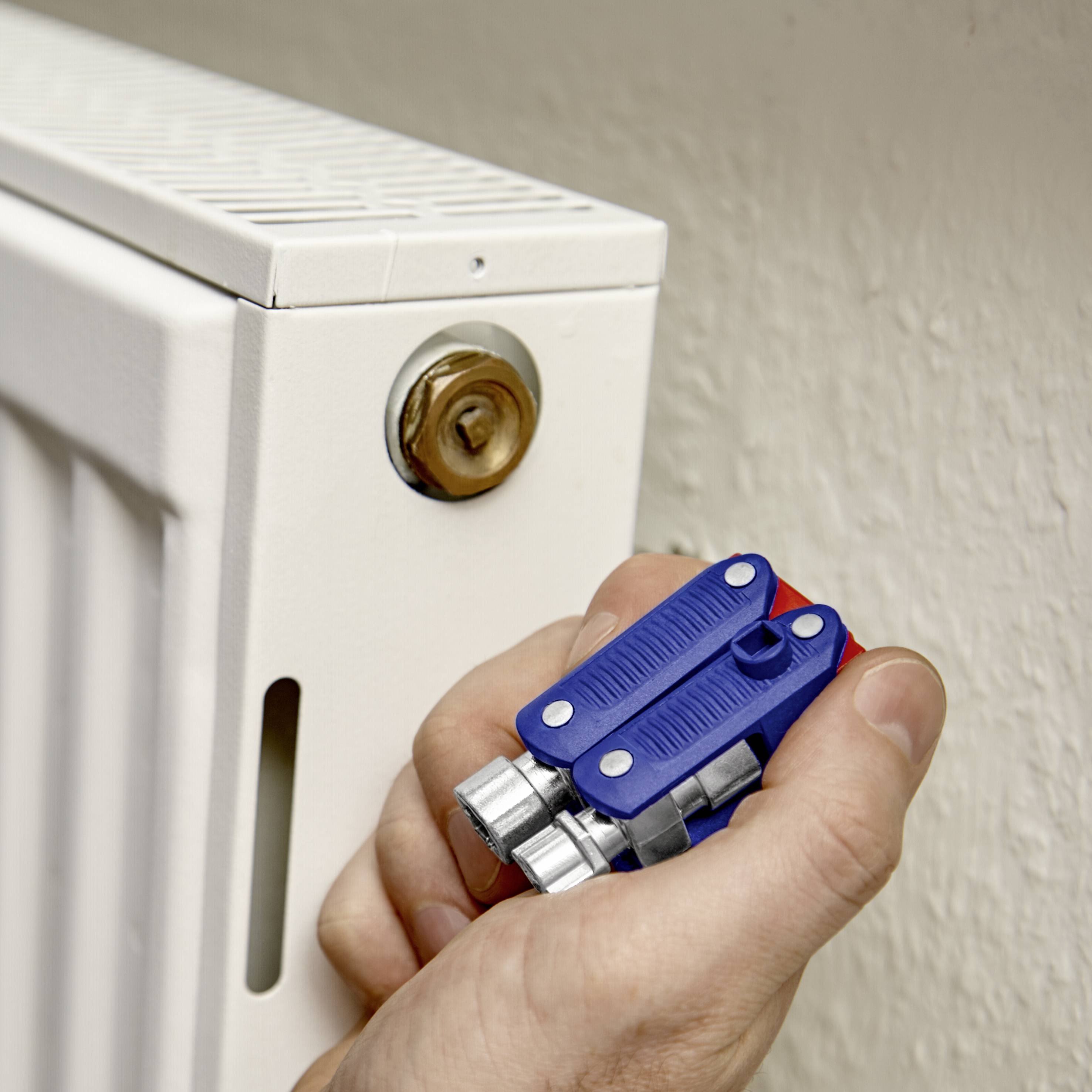 Une main tient une clé de radiateur devant un radiateur blanc. La clé est positionnée sur la valve pour purger l'air.