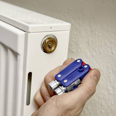 Une main tient une clé de radiateur devant un radiateur blanc. La clé est positionnée sur la valve pour purger l'air.