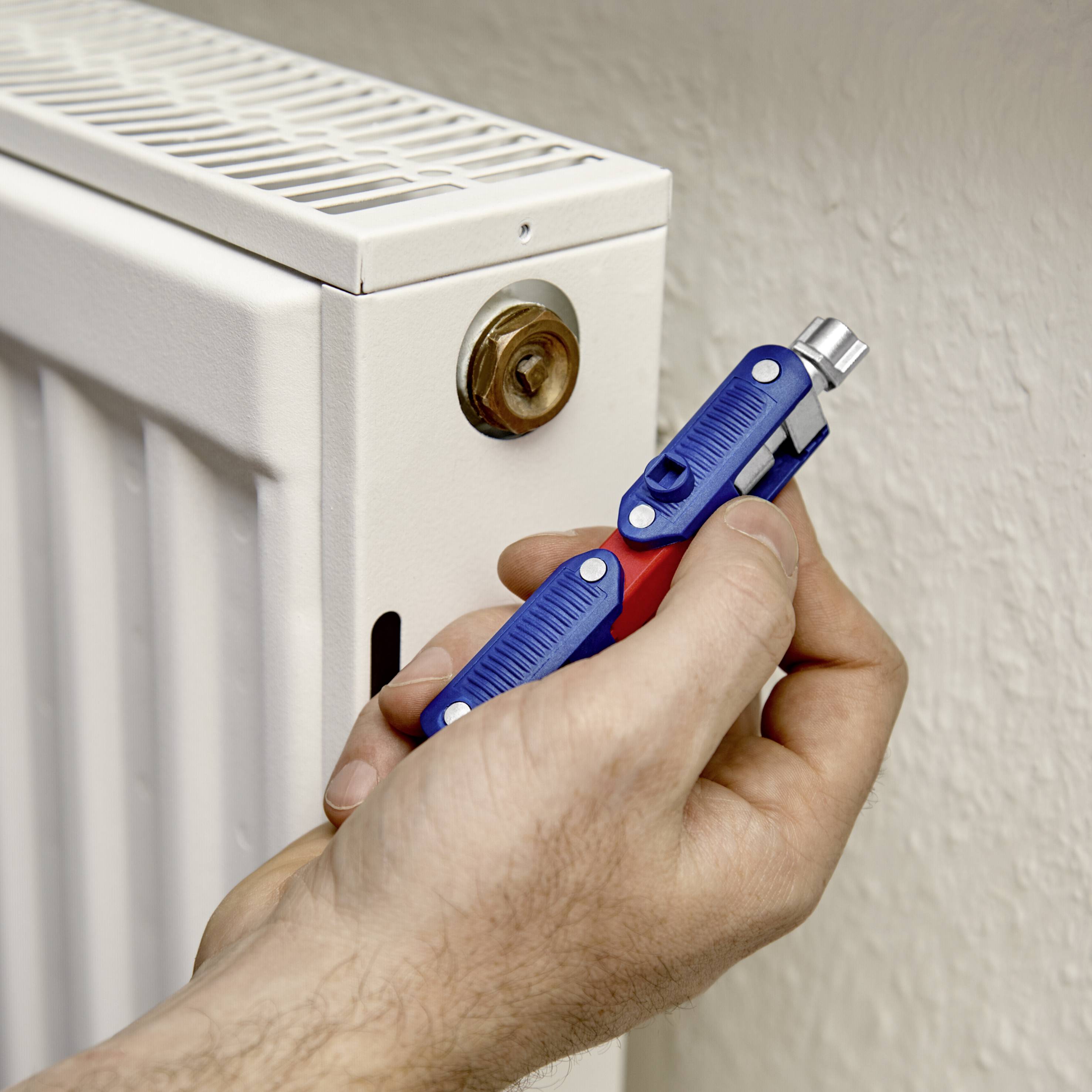 Une main purge un radiateur avec une clé de purge. Le radiateur est monté au mur et peint en blanc.