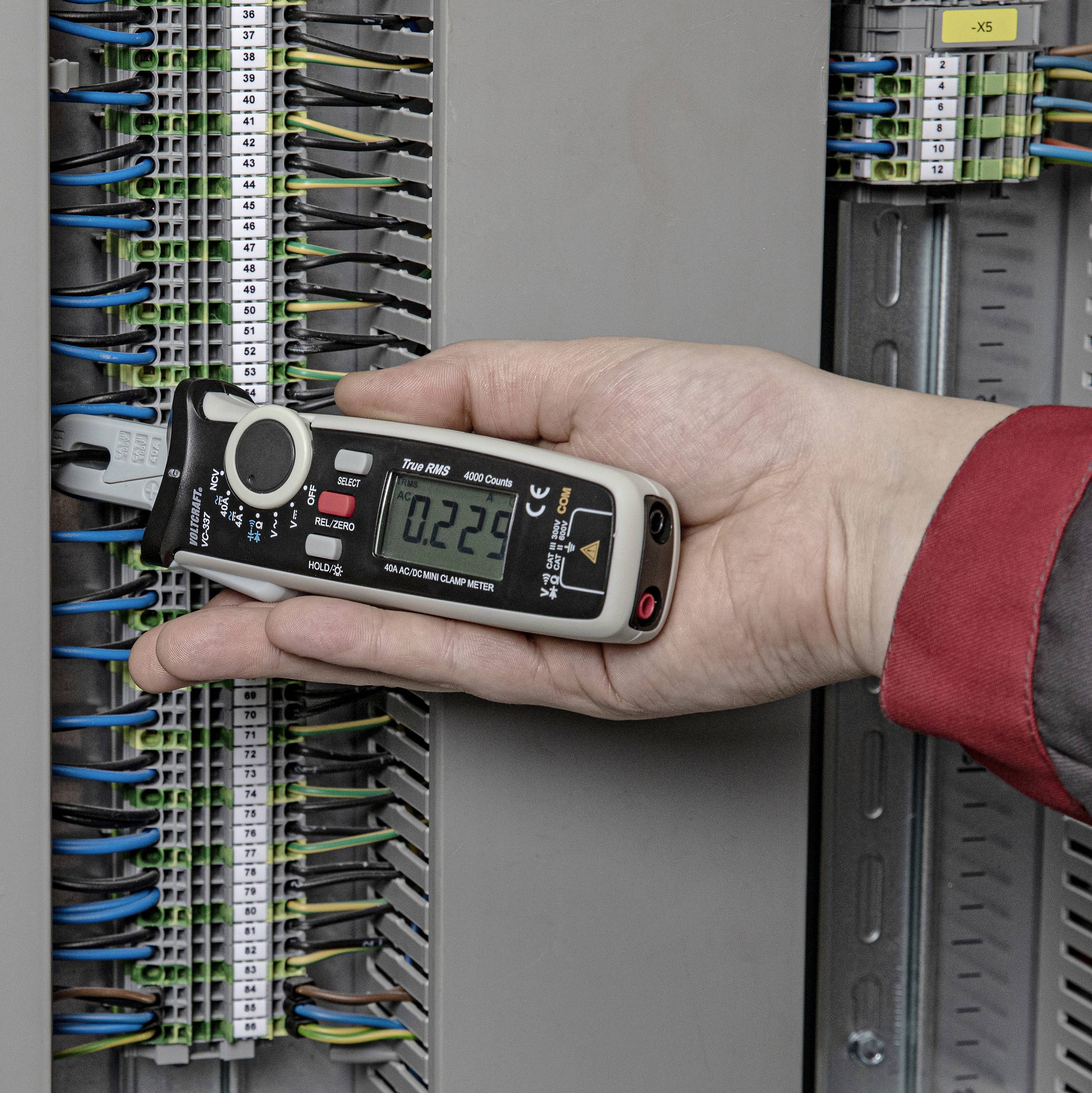 Une main tient un multimètre connecté à un armoire électrique ouverte afin de mesurer la tension électrique.