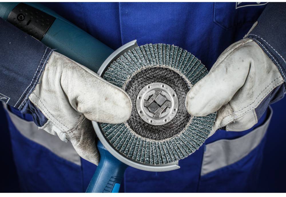 Un ouvrier en tenue bleue et portant des gants tient un disque abrasif sur une meuleuse d'angle, prêt à travailler le métal.