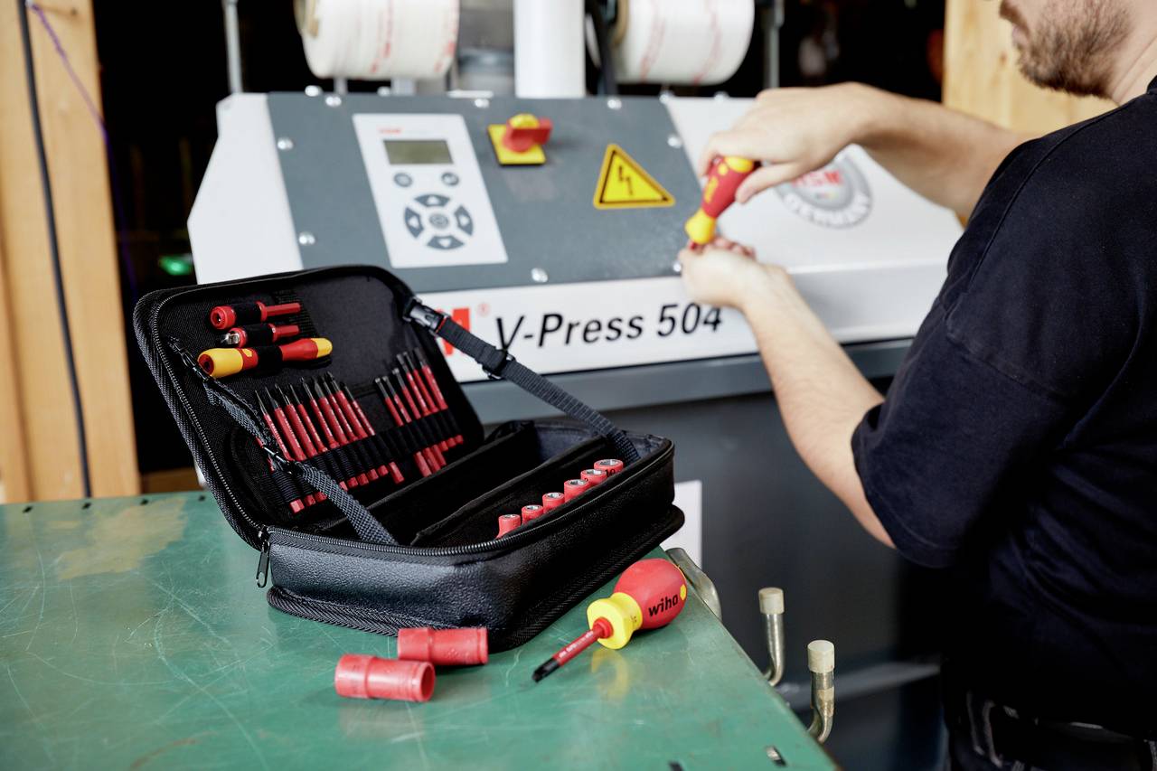 Un technicien travaille sur une machine, équipé d'une mallette à outils remplie de tournevis. Un panneau de commande de la machine est visible en arrière-plan.