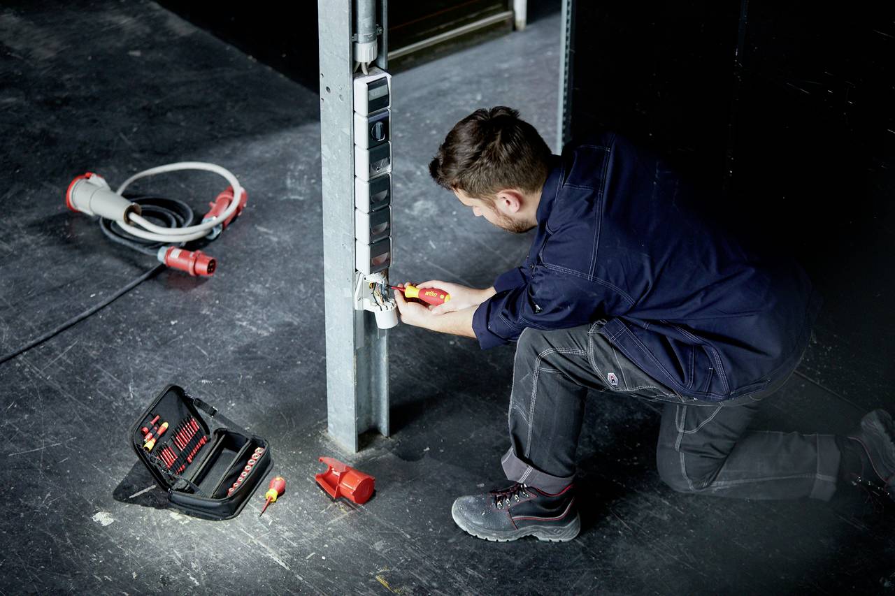 Un homme en tenue de travail répare une prise électrique dans un environnement industriel. Une boîte à outils et des câbles sont posés au sol.