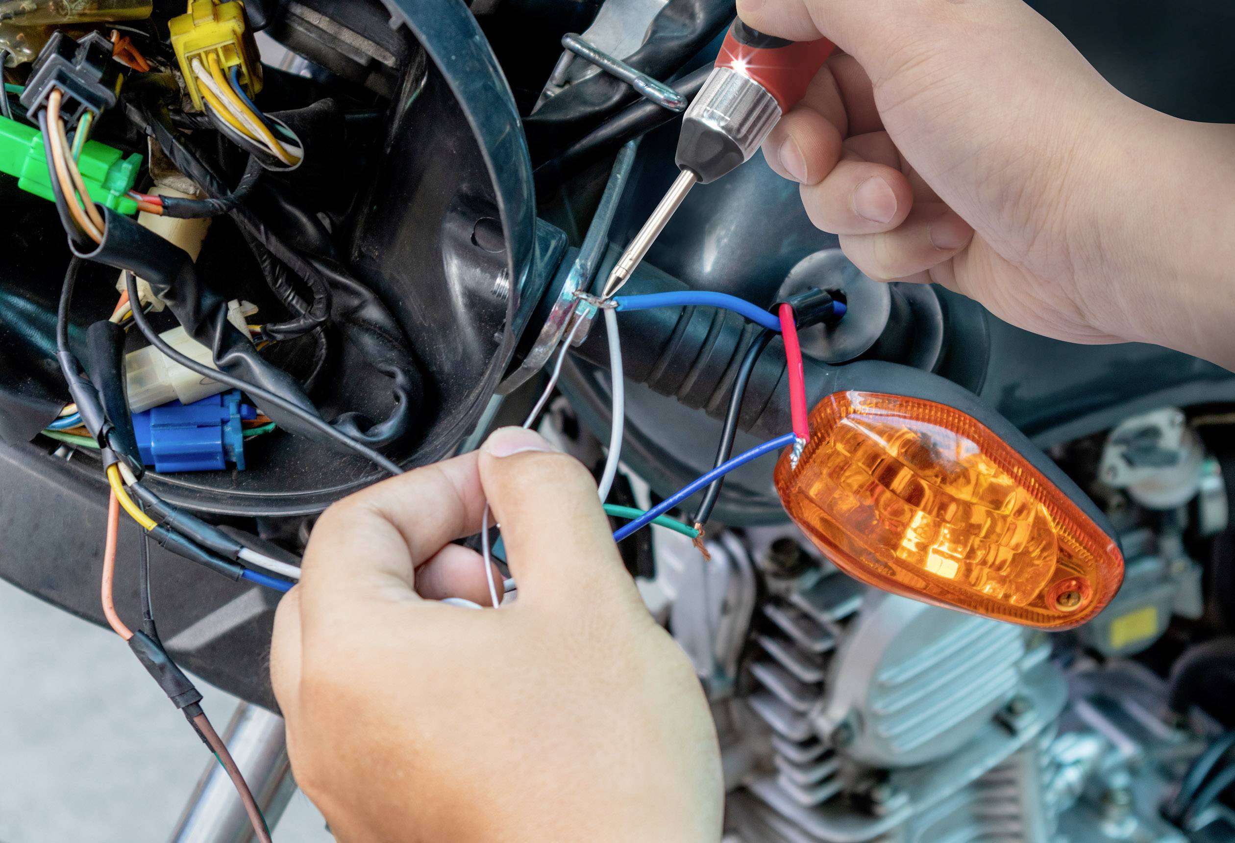 Une personne utilise un tournevis pour connecter des fils sur une moto. Un voyant clignotant orange est visible.
