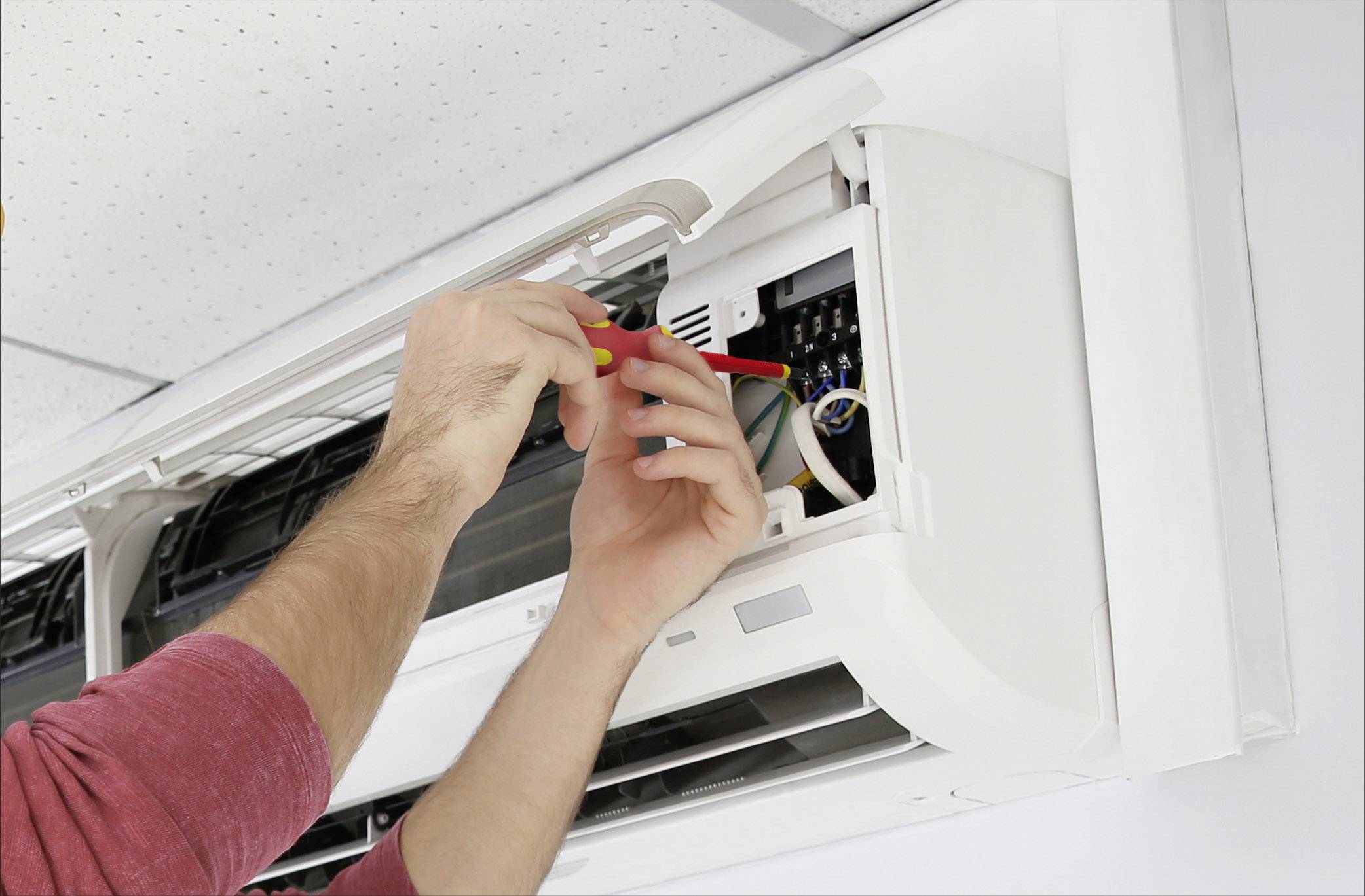 Une personne répare un climatiseur fixé au mur. Des mains tenant un tournevis travaillent à l'intérieur de l'appareil.