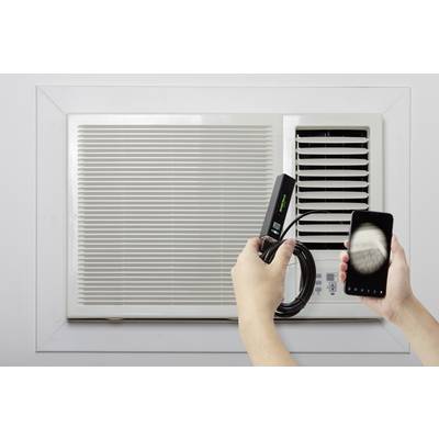Une personne tient un smartphone et un appareil câblé près d'un climatiseur. L'action suggère une connexion ou une mesure.