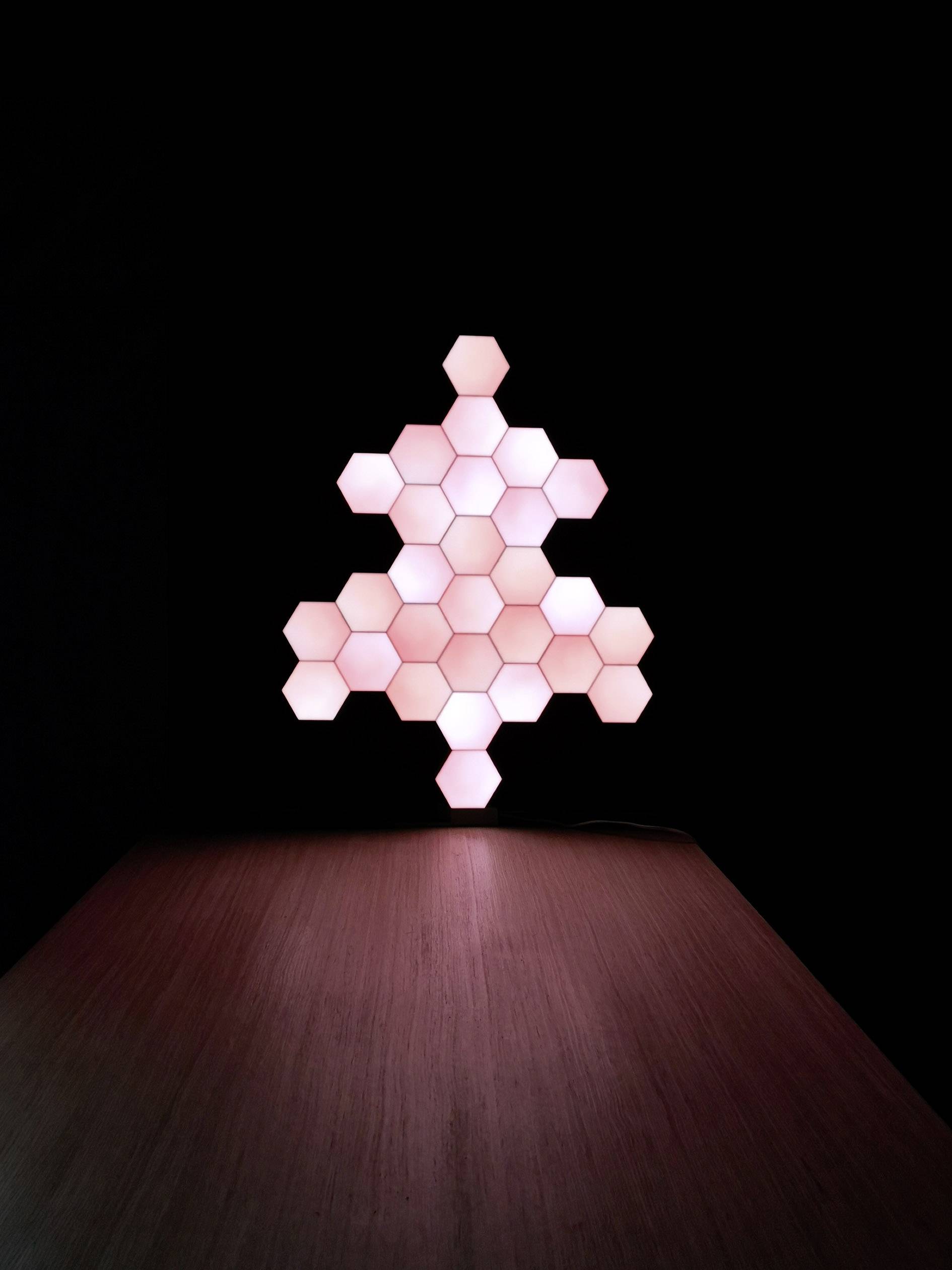 Panneaux lumineux en forme de sapin, composés de modules hexagonaux, placés sur une table et produisant une lumière douce et indirecte dans un environnement sombre.