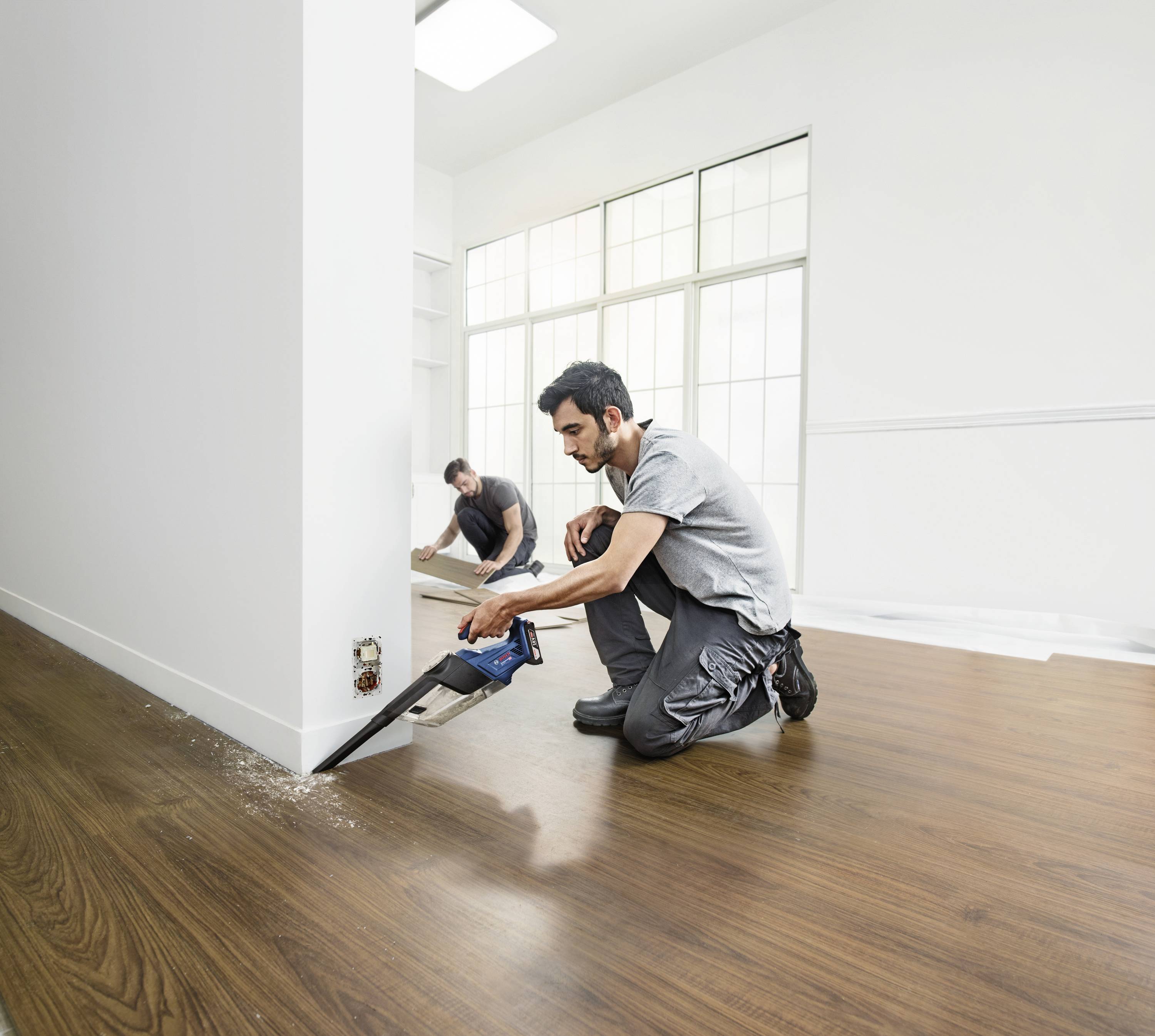 Deux hommes posent un parquet en bois dans une pièce lumineuse. L'homme devant scie avec une scie électrique, celui derrière est à genoux.