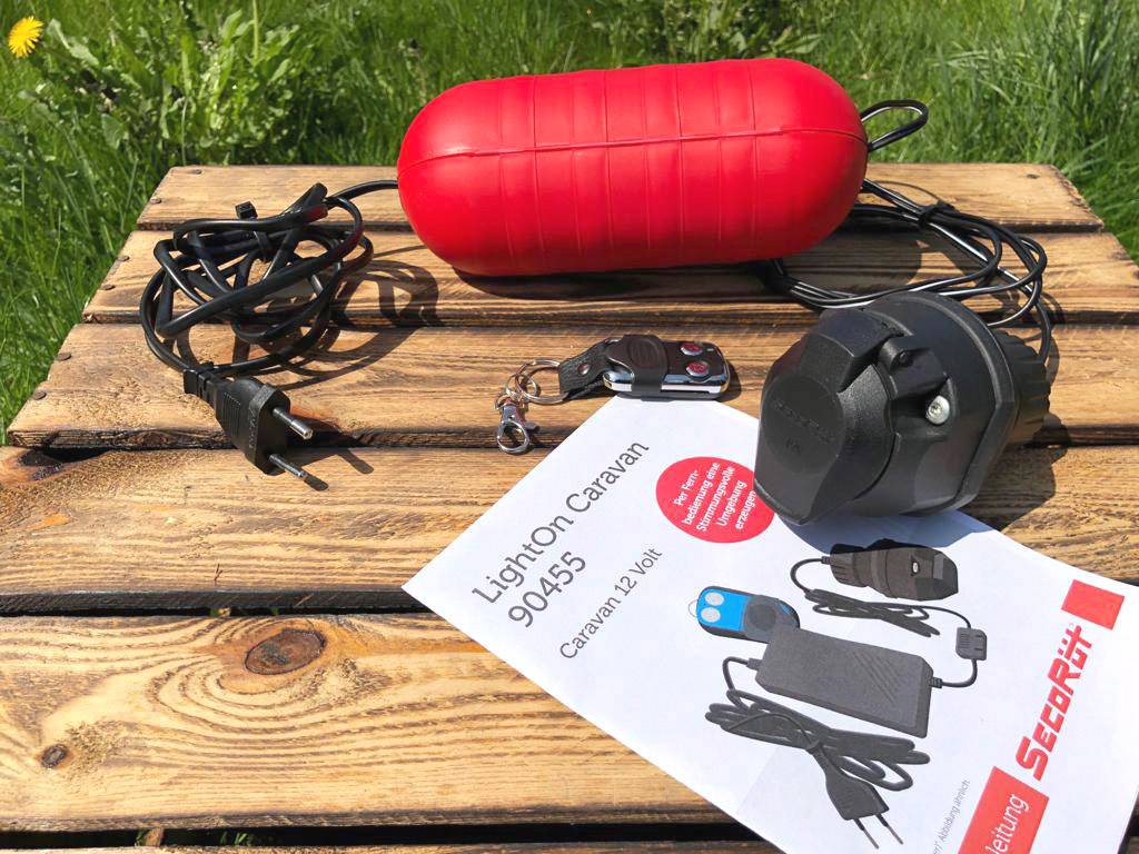 Un appareil rouge avec des câbles, un adaptateur et une télécommande est posé sur une table en bois. Un papier portant l'inscription « LightOn Caravan 90455, Caravan 12 Volts » est visible à côté.