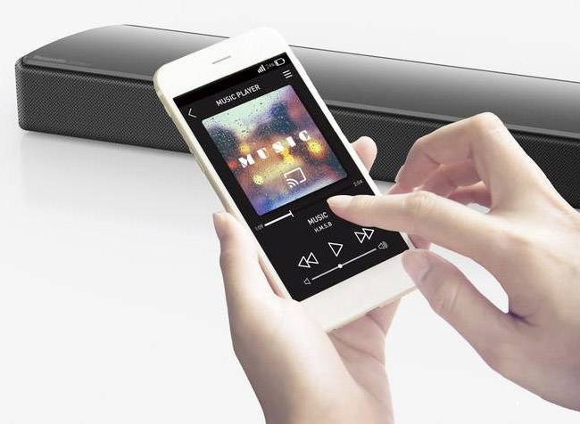 Une personne utilise un lecteur de musique sur smartphone ; des enceintes sont visibles en arrière-plan ; l'écran affiche la pochette de l'album et les commandes de lecture.