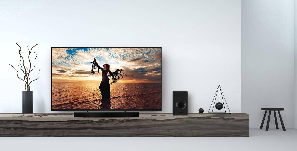 Téléviseur sur une étagère en bois montrant une femme au chapeau sur une plage au coucher de soleil. À côté, des enceintes et une décoration minimaliste.