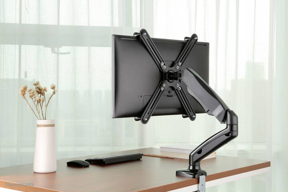 Un écran d'ordinateur est monté sur un bras de moniteur ajustable posé sur une table en bois. À côté de l'écran se trouvent un clavier, une souris et un vase contenant des fleurs séchées.