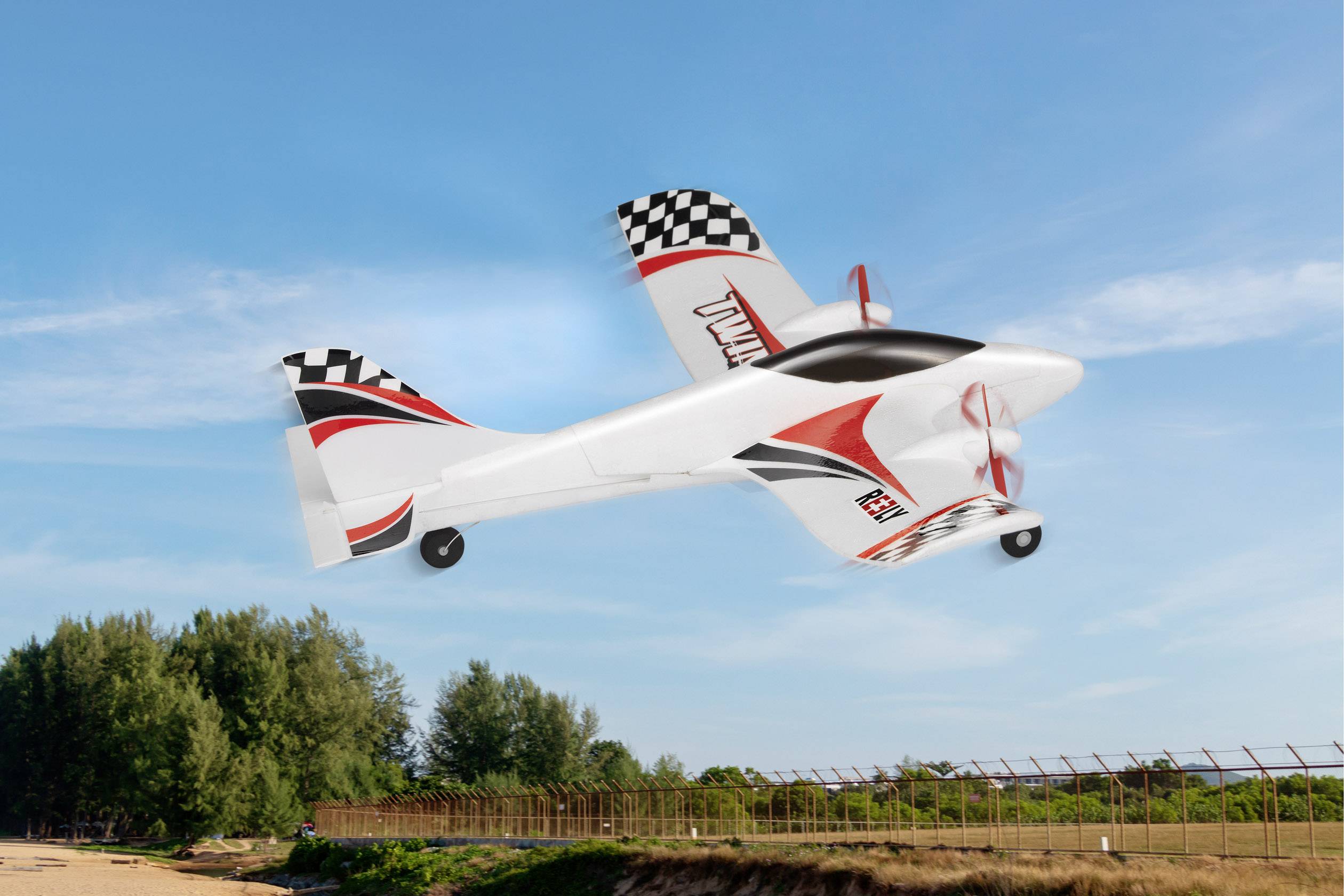 Montre un avion de sport télécommandé en vol au-dessus d'un champ, avec un ciel bleu et des arbres en arrière-plan.